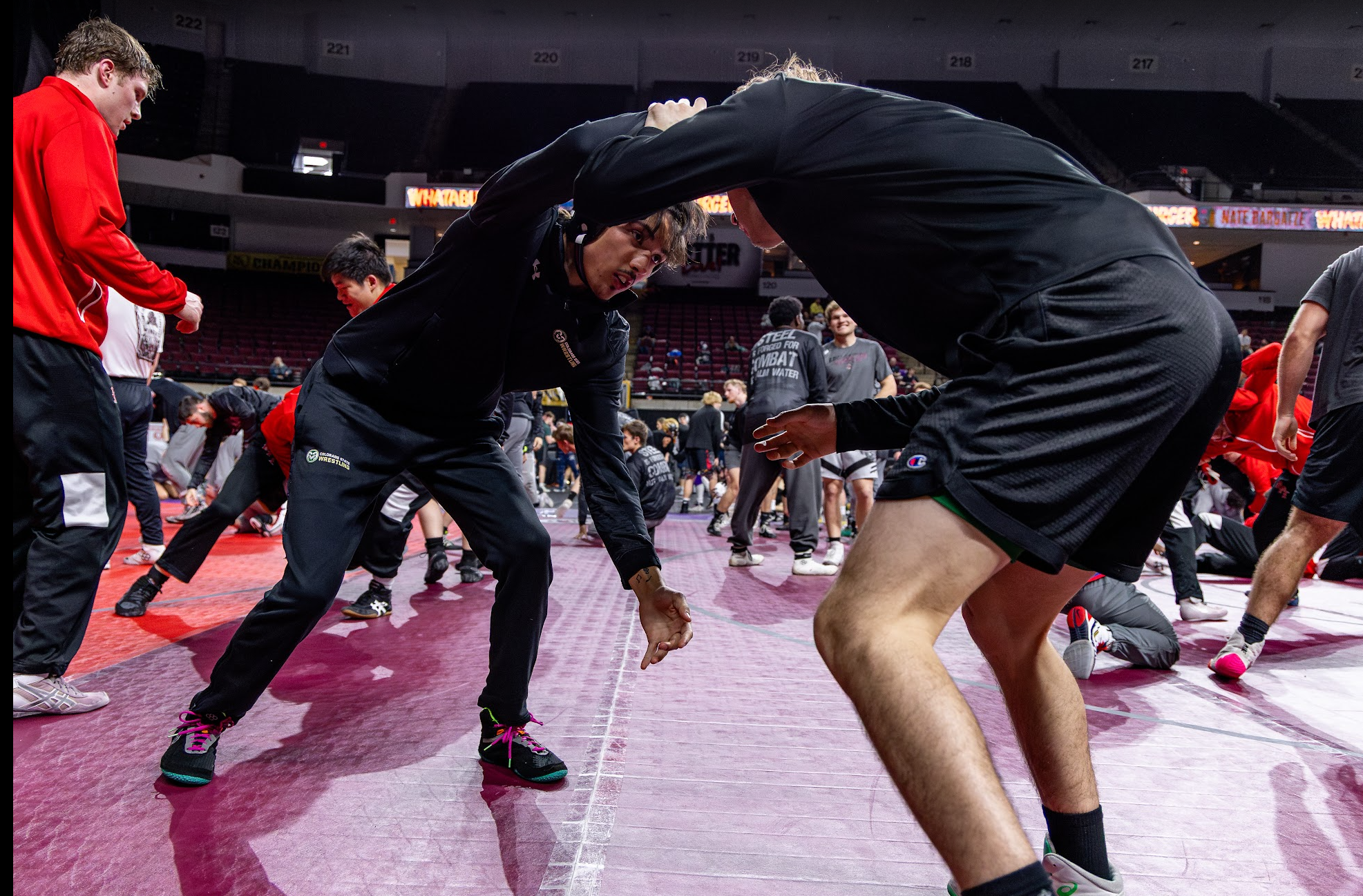 Wrestlers practicing on the mat in an arena filled with other wrestlers and spectators.