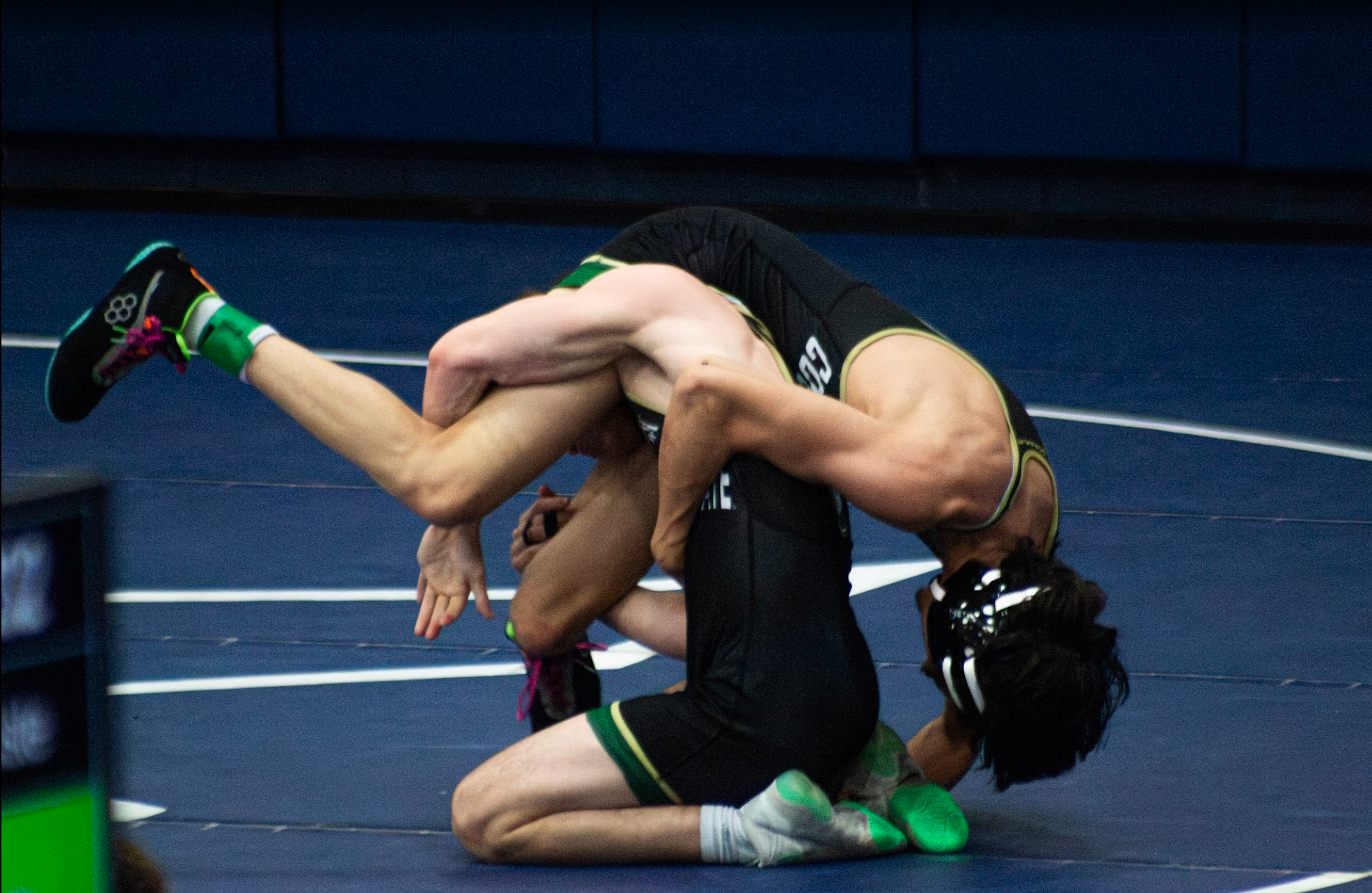 Two wrestlers engaged in a match on a blue wrestling mat, with one wrestler on top in a dominant position and the other underneath, both wearing black singlets and athletic shoes.