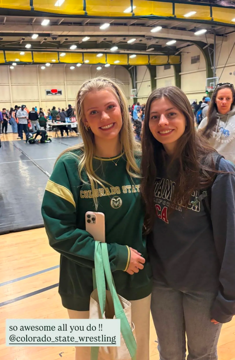 Two young women smiling at a wrestling event inside a gymnasium. One is wearing a Colorado State sweatshirt and the other a Cal State Dominguez Hills hoodie. In the background, there are people, tables, and a scoreboard.