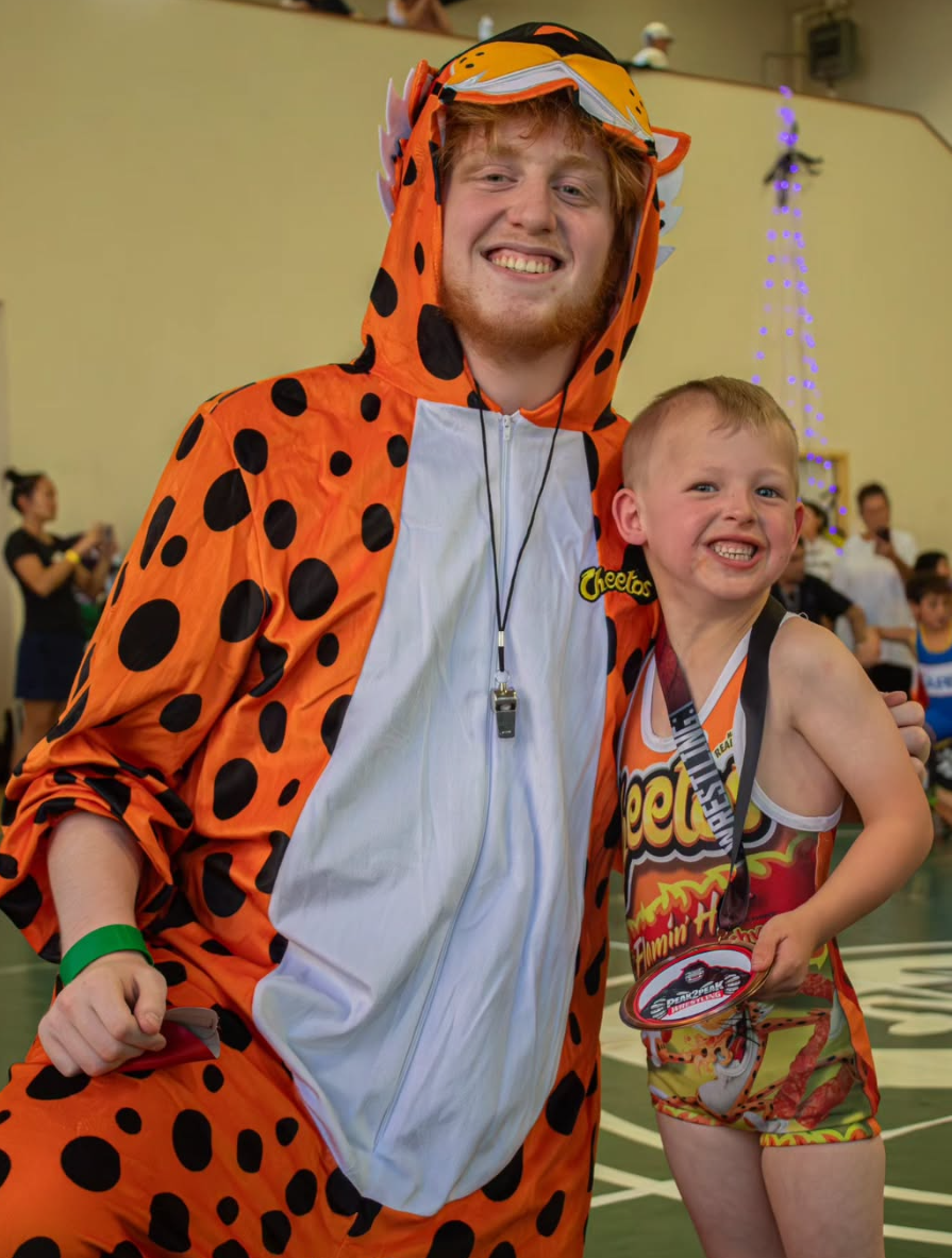 A man dressed in a cheetah costume and a child in a sports outfit with a Cheez-It logo, smiling and posing together at an indoor event, likely a sports or costume party.