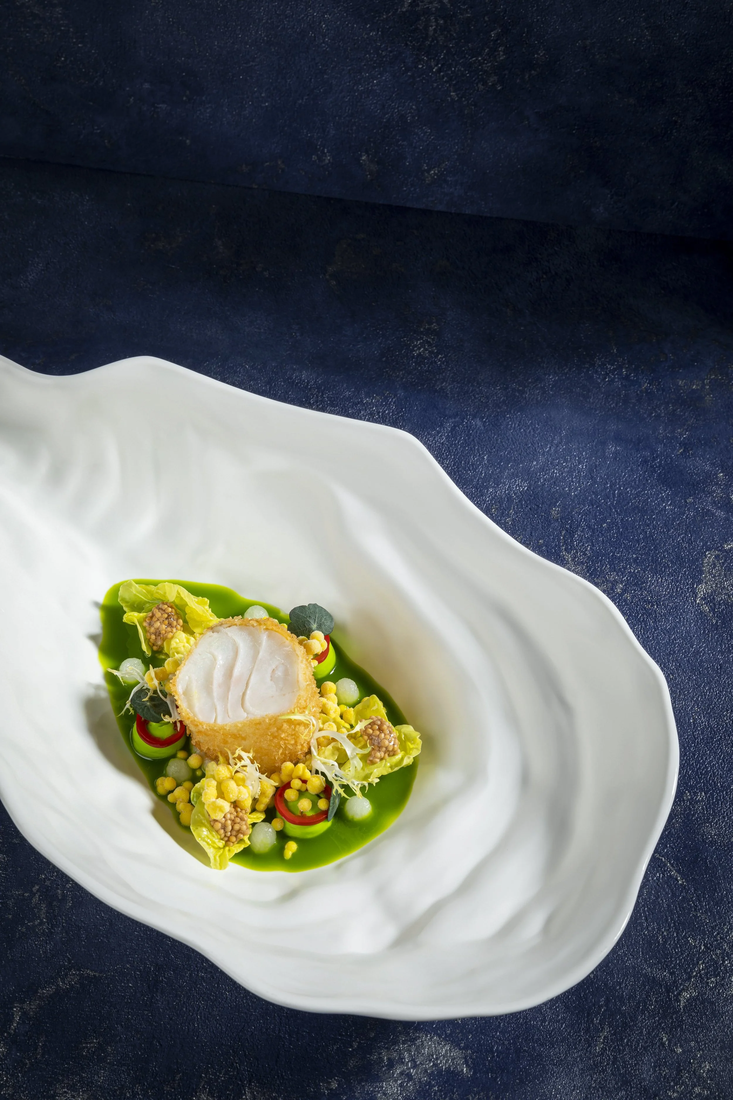 Gedünstetes Hähnchenfleisch in knuspriger Panade mit grünen und gelben Gemüsesorten auf grünlicher Soße, serviert in einer weißen geschwungenen Schale.