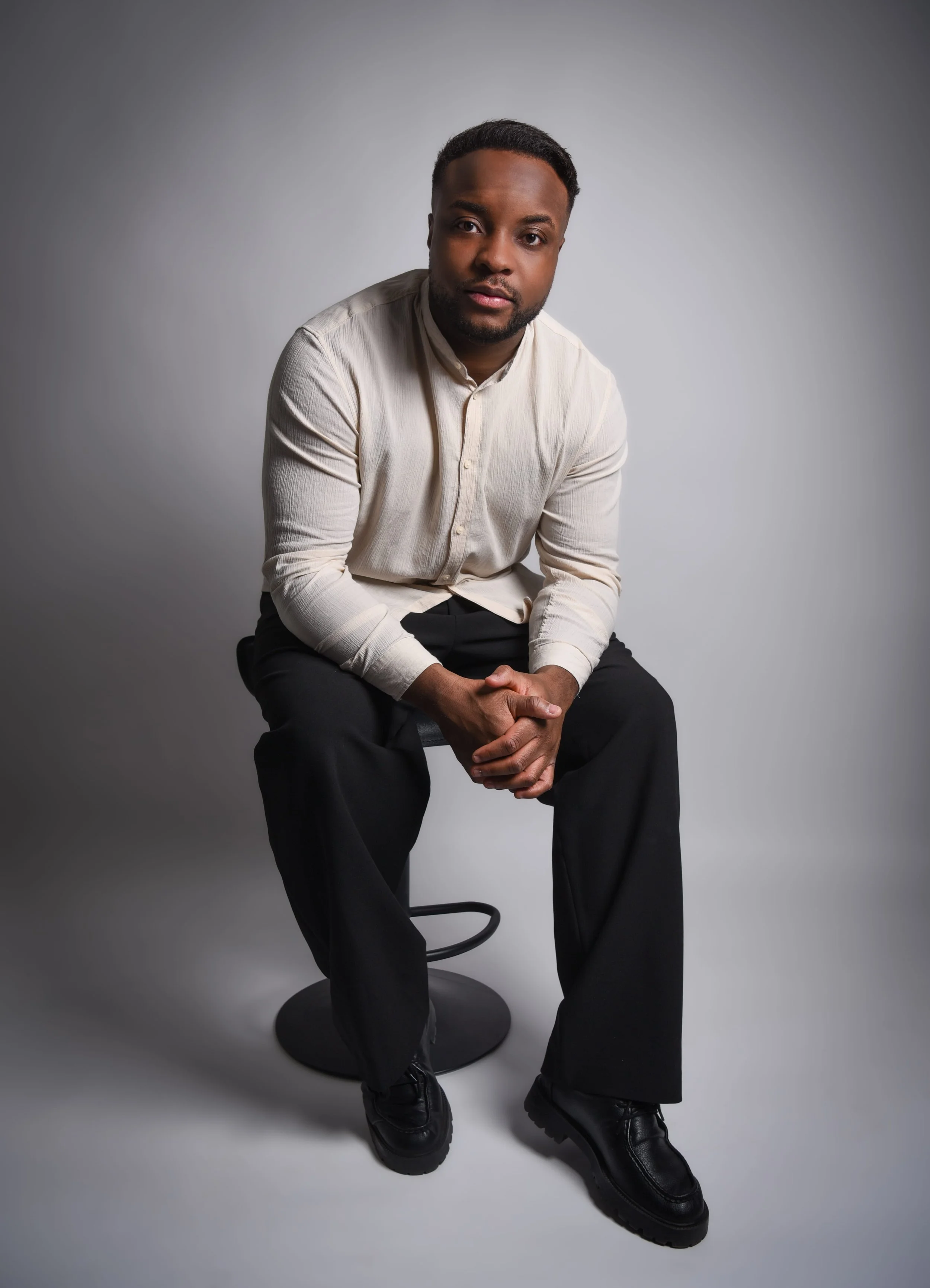 Ein Mann sitzt auf einem schwarzen Hocker vor grauem Hintergrund, trägt eine cremefarbene Hemd und schwarze Hosen.