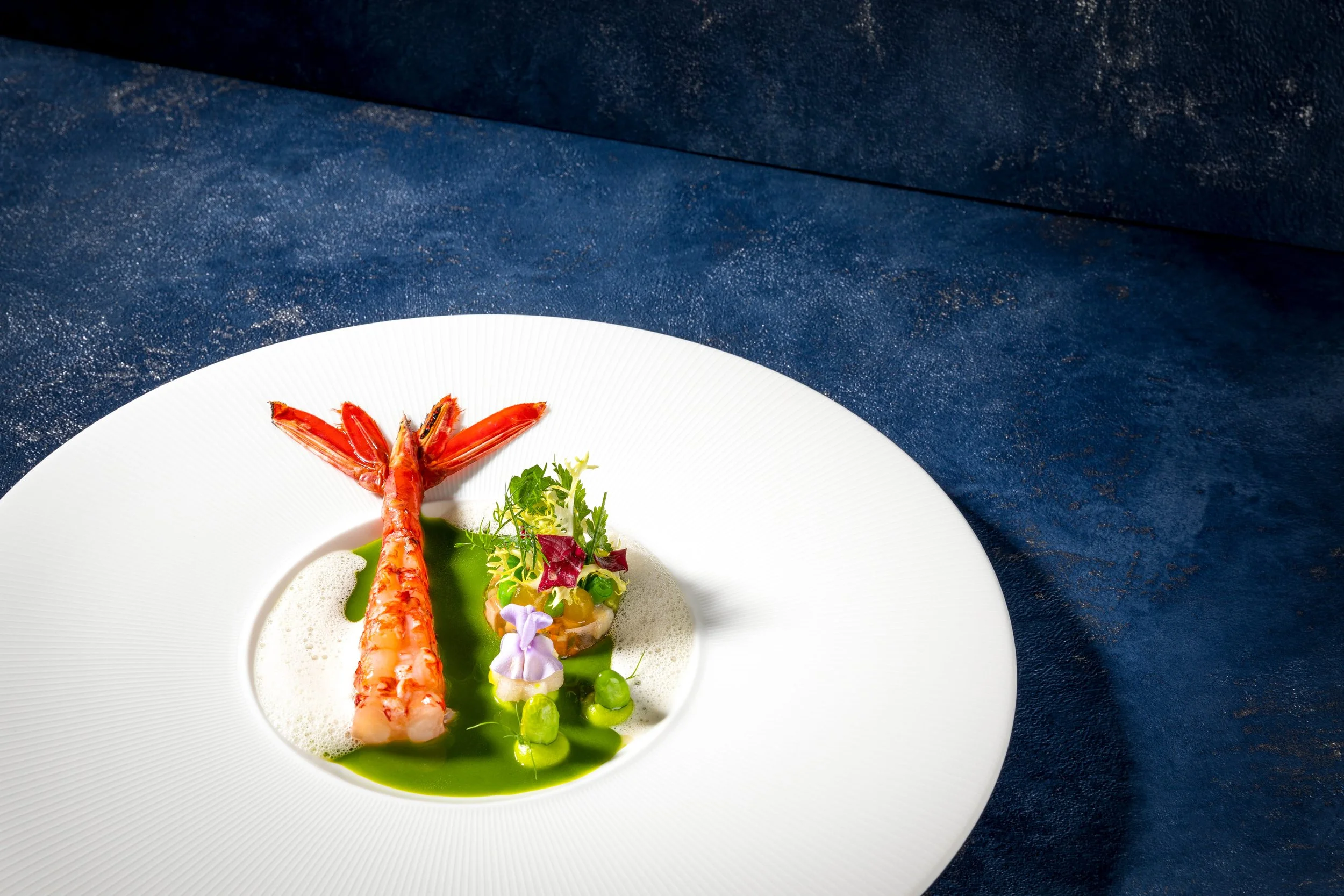 Elegantes Gericht mit Garnelen, grünem Sauce, Gemüse, Blüten und Microgreens auf weißem Teller auf dunkelblauem Hintergrund.