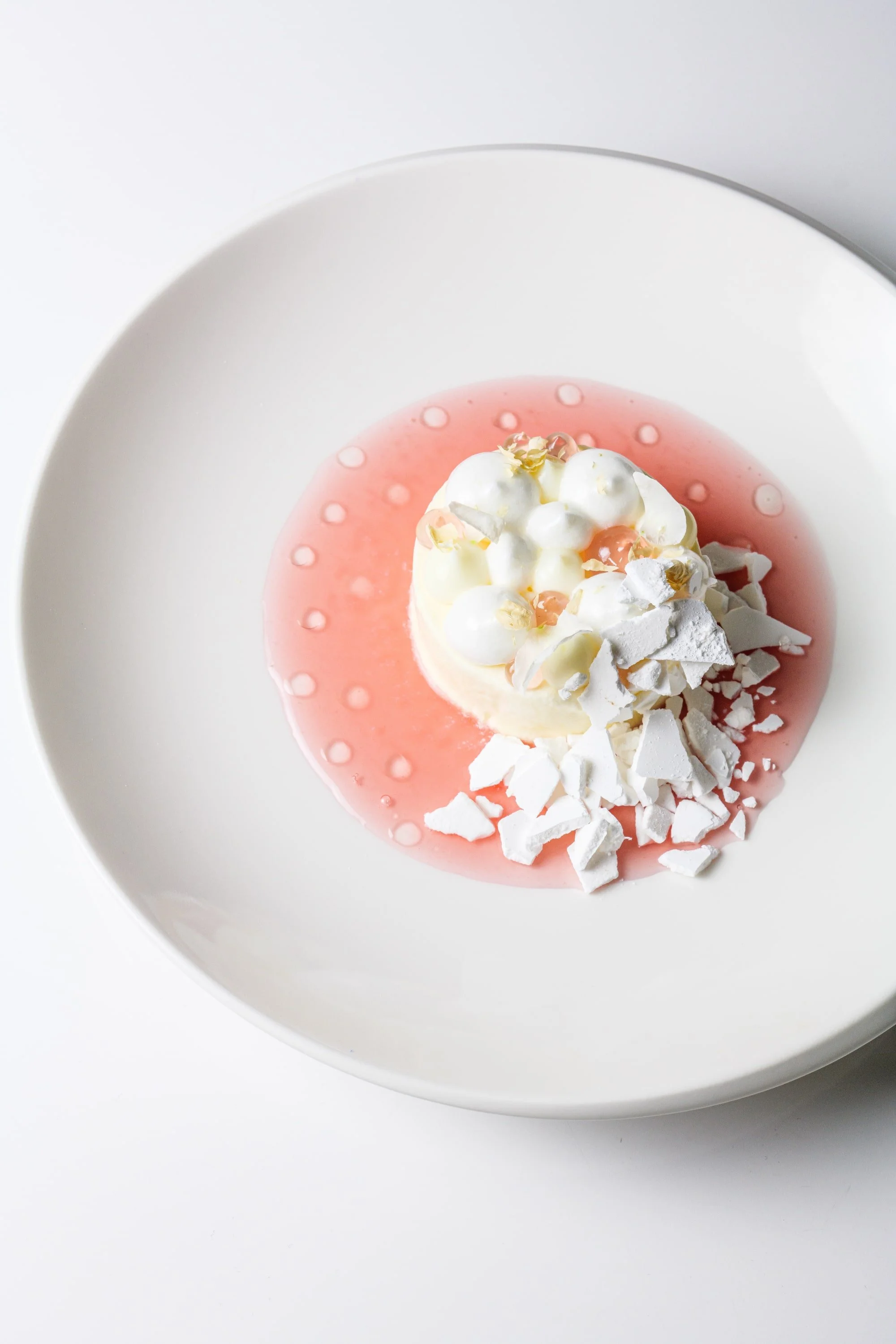 Ein Dessert mit weißer Schokoladenmousse, umgeben von rosa Gelee, dekoriert mit weißen Schokoladenstücken, kleinen weißen Bällchen und goldenen Essbaren Dekorationen auf einem weißen Teller.