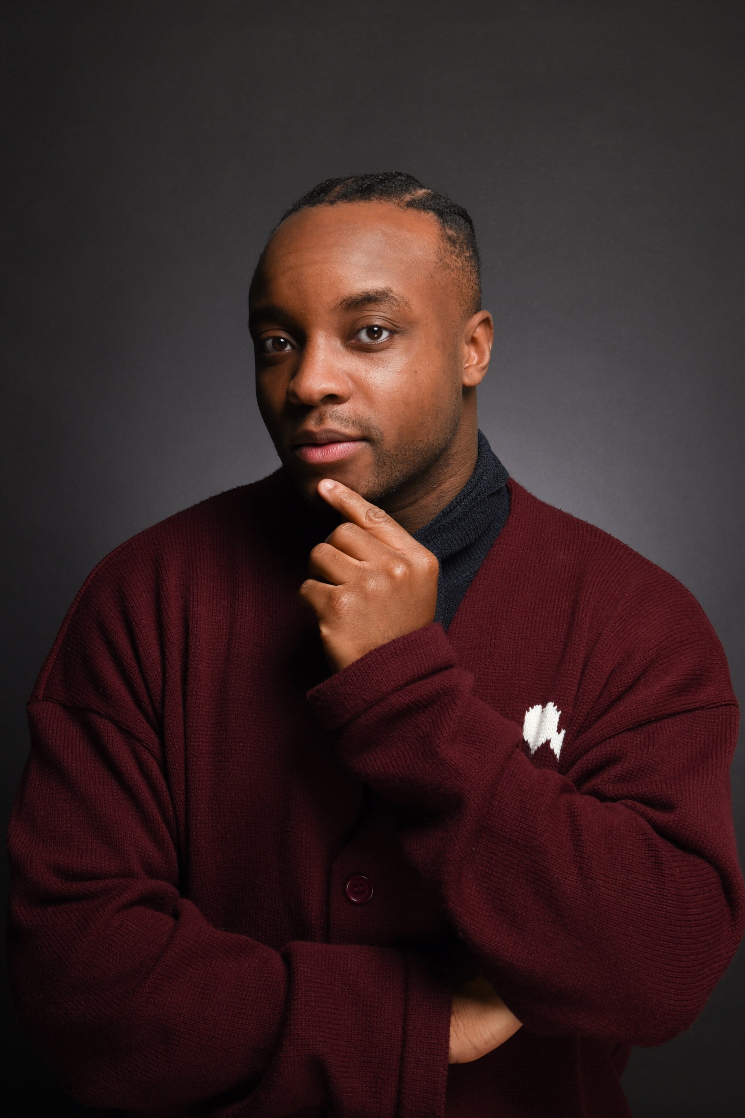 Porträt eines jungen Mannes mit dunkler Haut, schwarzen Haaren, in einem dunkelroten Pullover und dunklem Hemd vor einem dunklen Hintergrund, der nachdenklich mit der Hand am Kinn posiert.