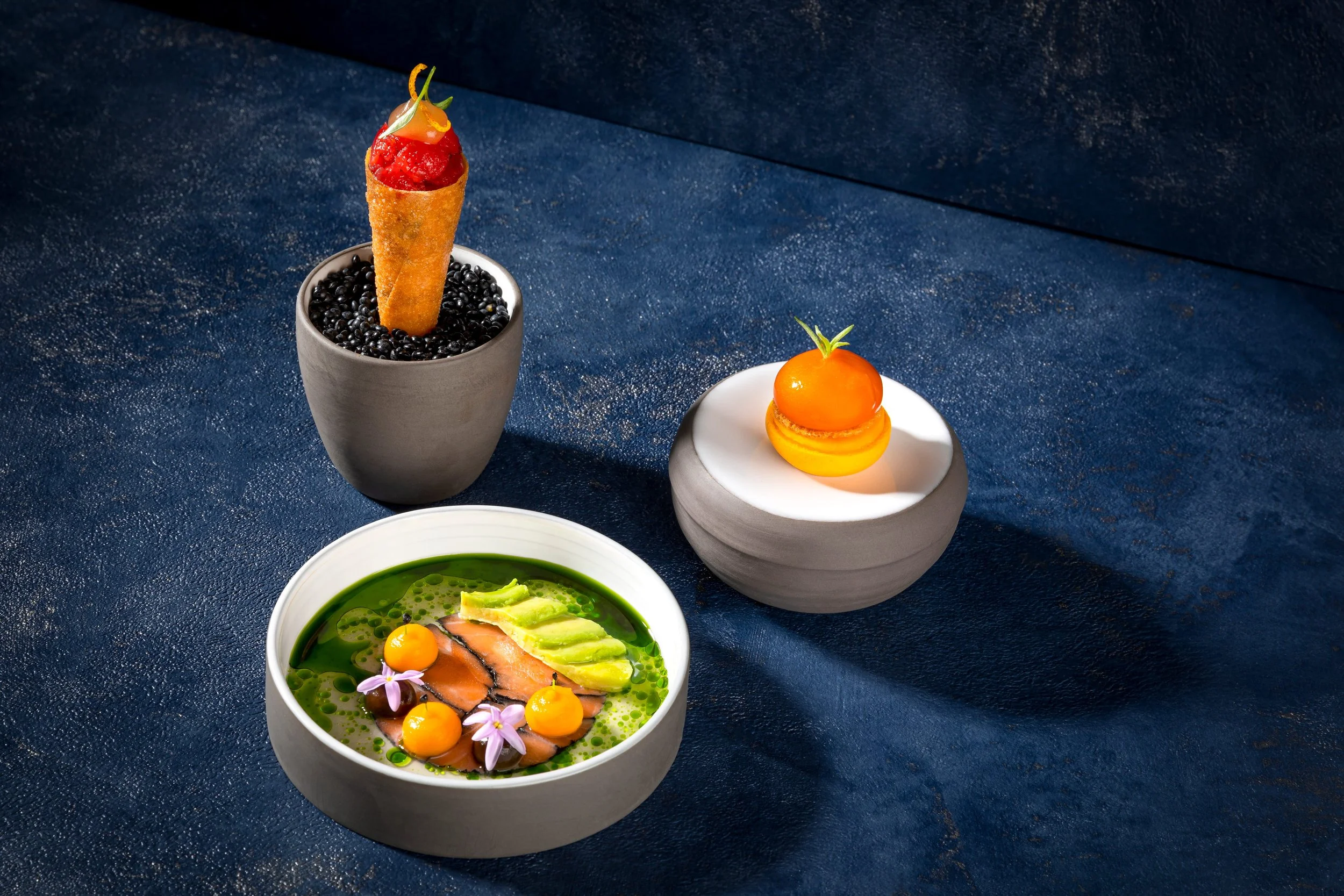 Drei kunstvoll zubereitete Gerichte auf schwarzen Oberflächen, inklusive einer Schale mit rotem Surimi in einer Waffel, einer Schale mit Orangencurry und Lachs, und einer kleinen Schale mit Tomate, Zitronen, und grünen Blätter.