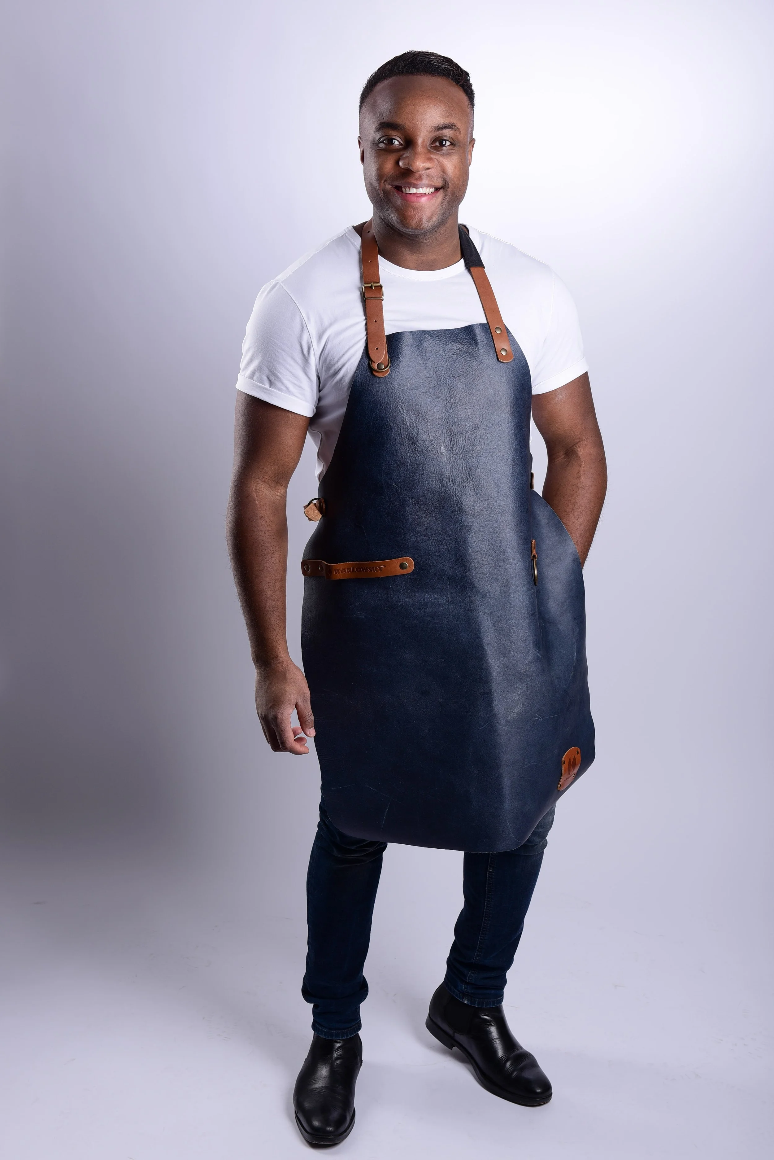 Ein lächelnder Mann trägt eine weiße T-Shirt und eine dunkle Lederschürze, steht vor einem hellgrauen Hintergrund.