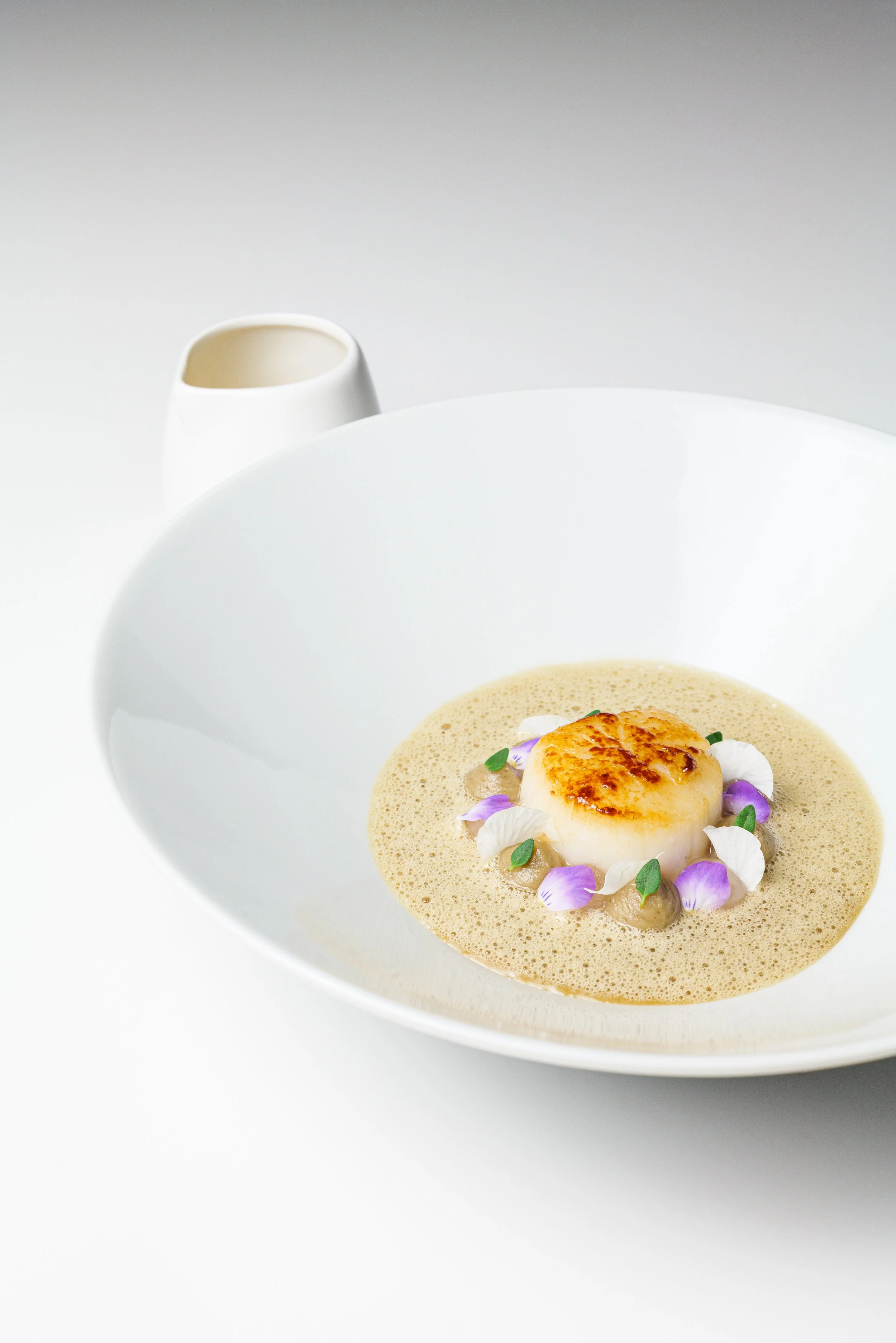 Feines Gericht mit einer gebratene Jakobsmuschel, verziert mit essbaren Blumen und kleinen Blättchen, in einer weißen Suppenschale auf weißem Hintergrund.