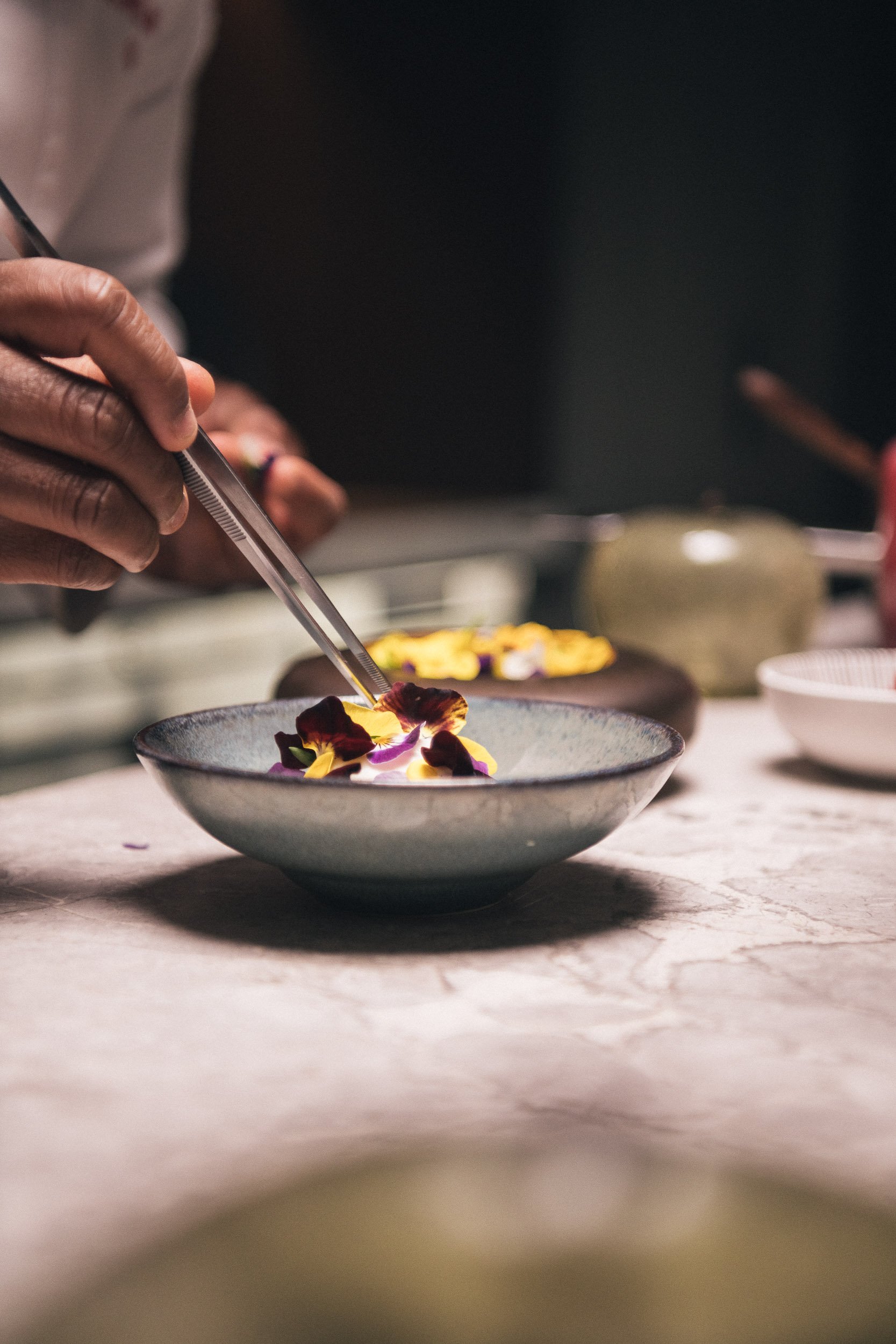 Person gebruikt pincetten, um essbare Blüten auf einen Teller zu legen.
