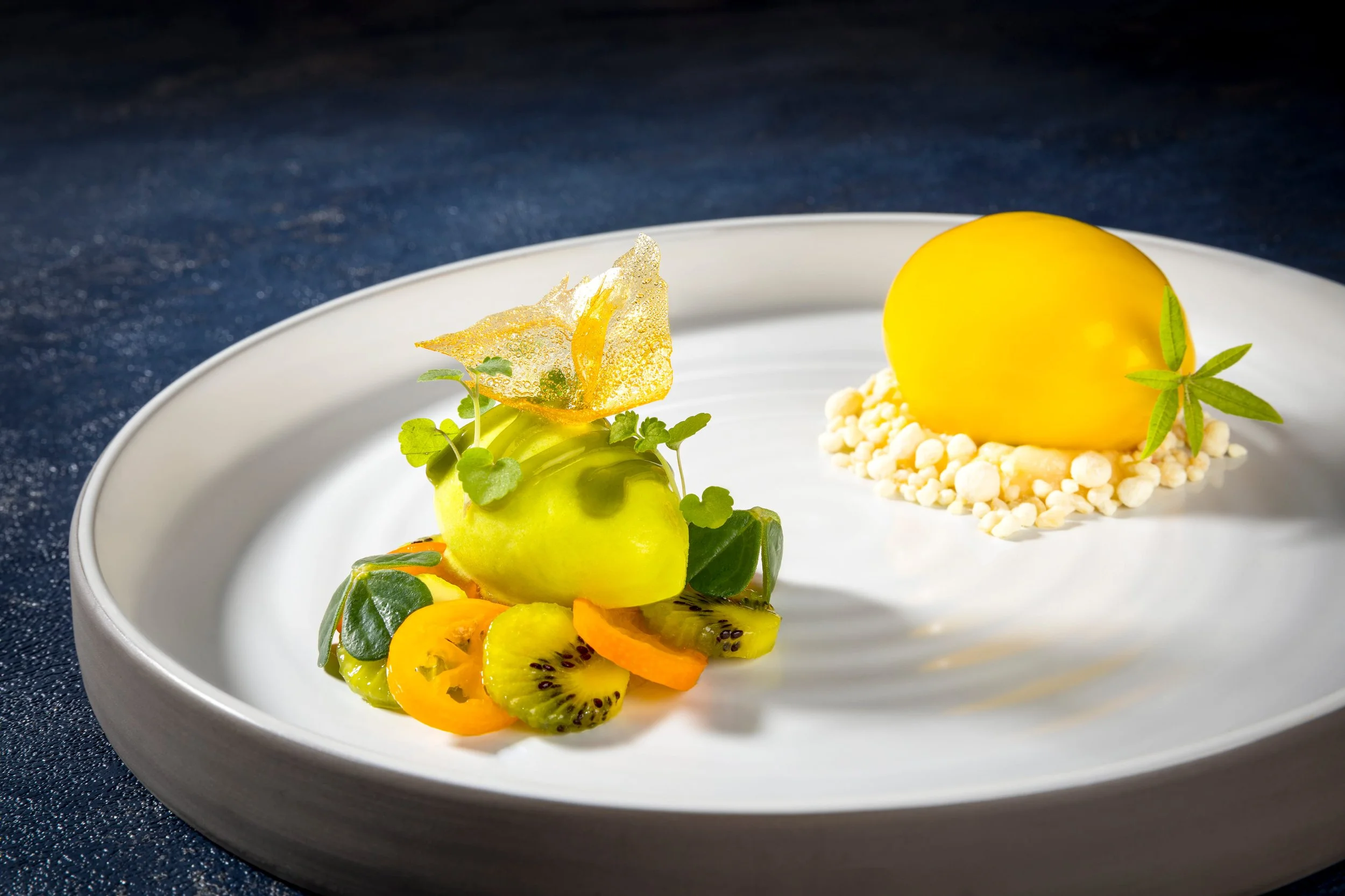 Ein Teller mit kunstvoll angerichteten Desserts, darunter eine gelbe, runde Süßigkeit und ein bunter Fruchtsalat mit Kiwi, Mango, Mango, Orange und Minzblättern auf einem weißen Teller.