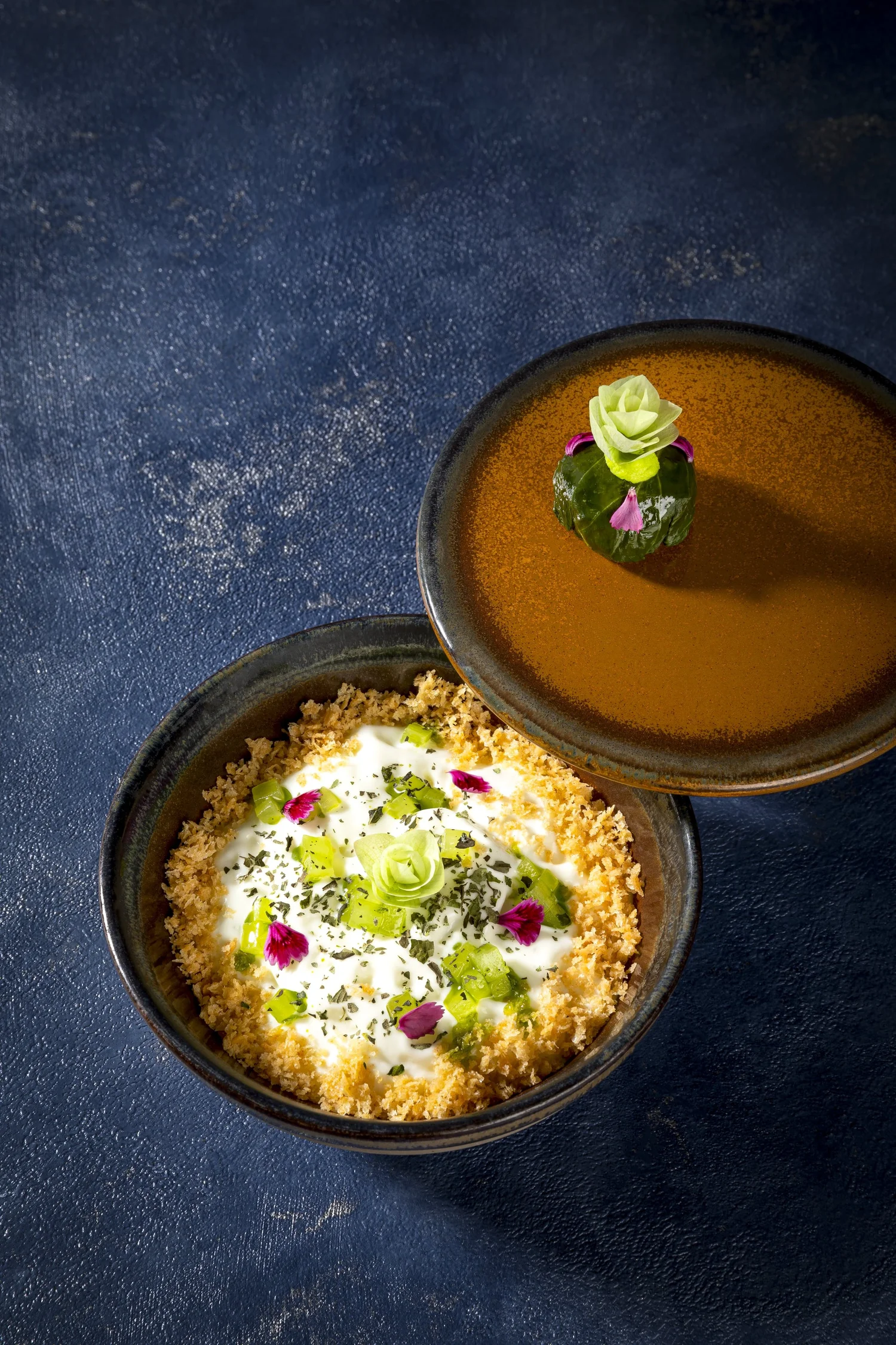 Zwei kleine Schüsseln mit chinesischer Suppe, dekoriert mit Frühlingszwiebeln, Kräutern und essbaren Blumen, auf einer dunklen Oberfläche.