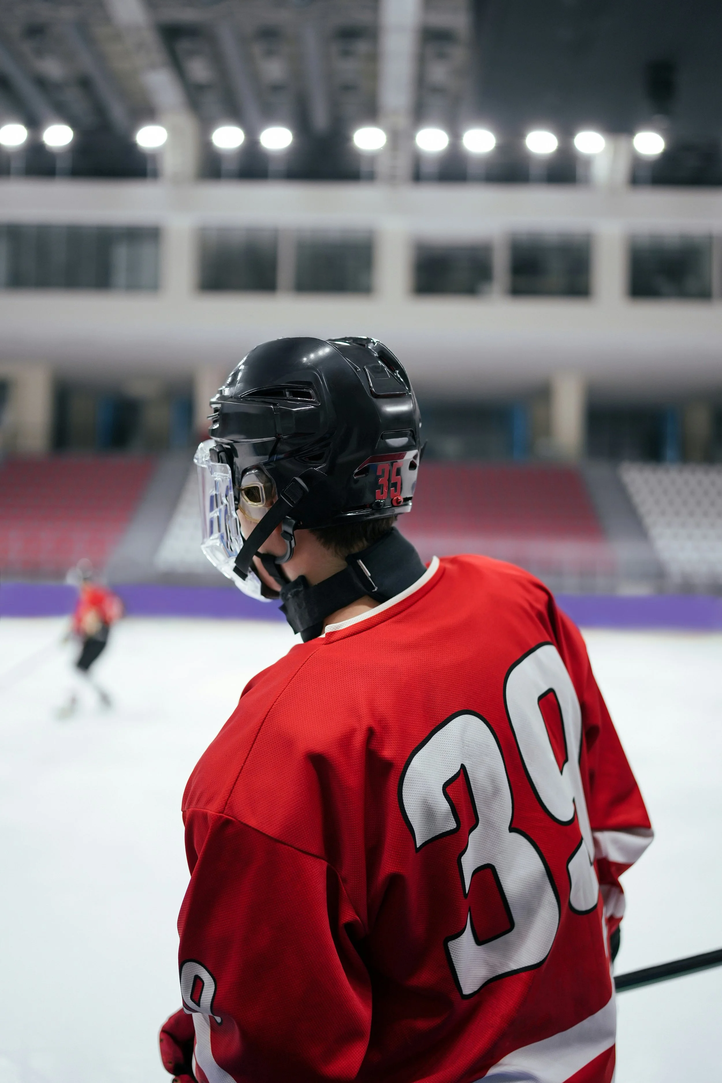 Ice hockey player wearing a black helmet with the number 35 and red jersey with the number 30 on the back, standing on an ice rink with other players in the background.