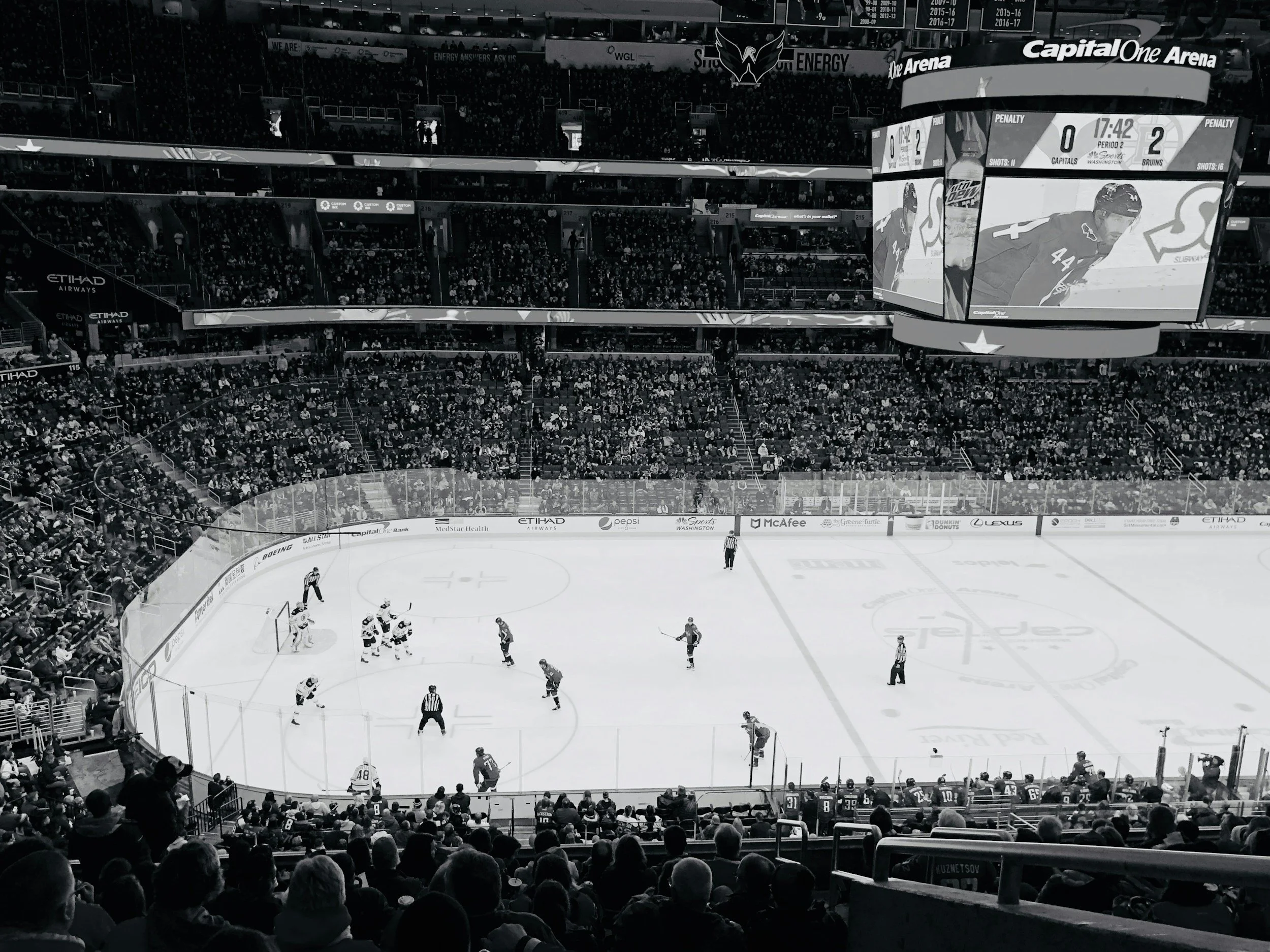 An ice hockey game is underway inside a large stadium filled with spectators. The scoreboard shows a score of 0-2 in the second period with 17 minutes and 42 seconds remaining. Players are on the ice, and the audience watches from the stands.