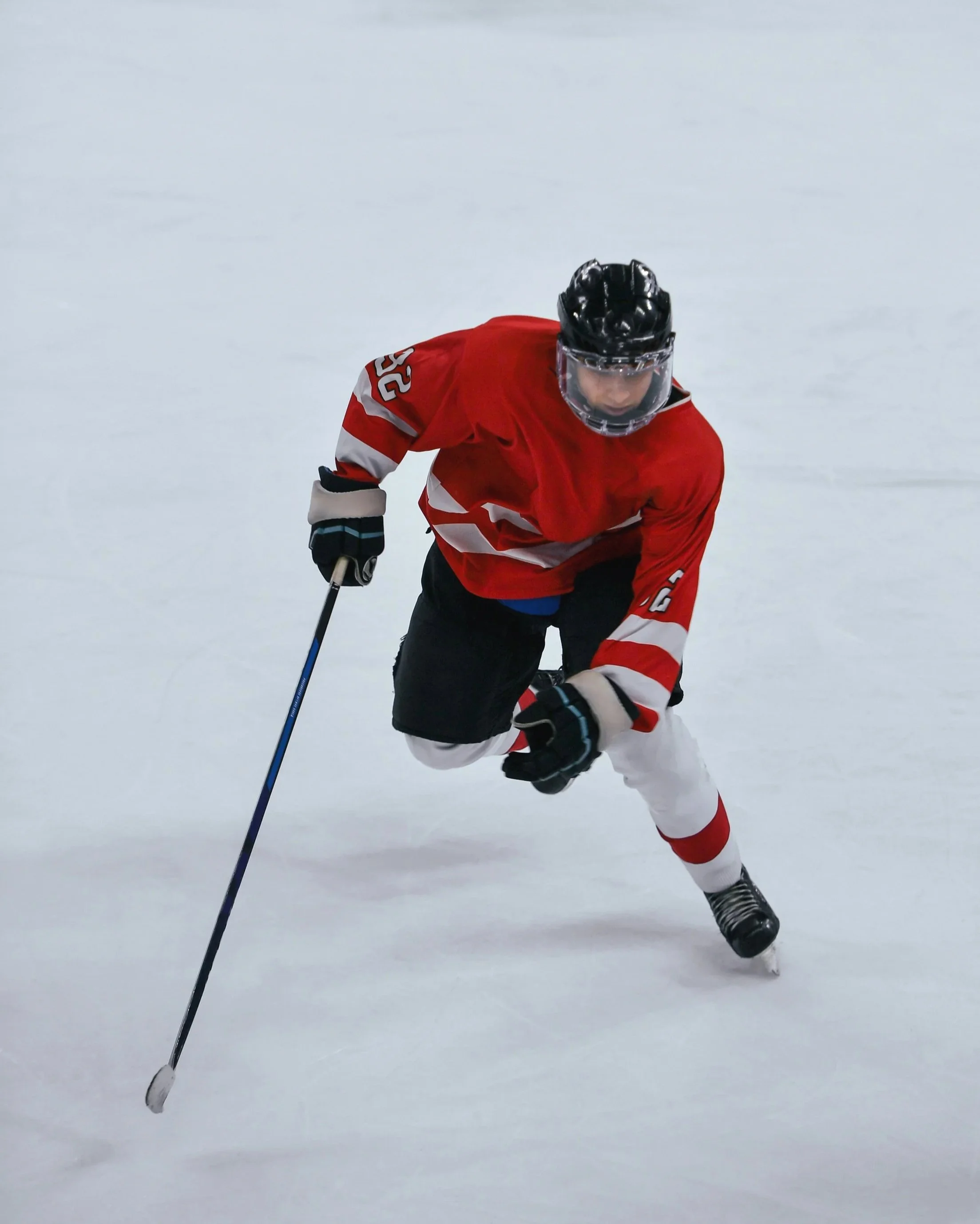 A hockey player in a red jersey with the number 23 on the sleeve, wearing a black helmet and gloves, skating on ice with a hockey stick, looking down towards the ice.