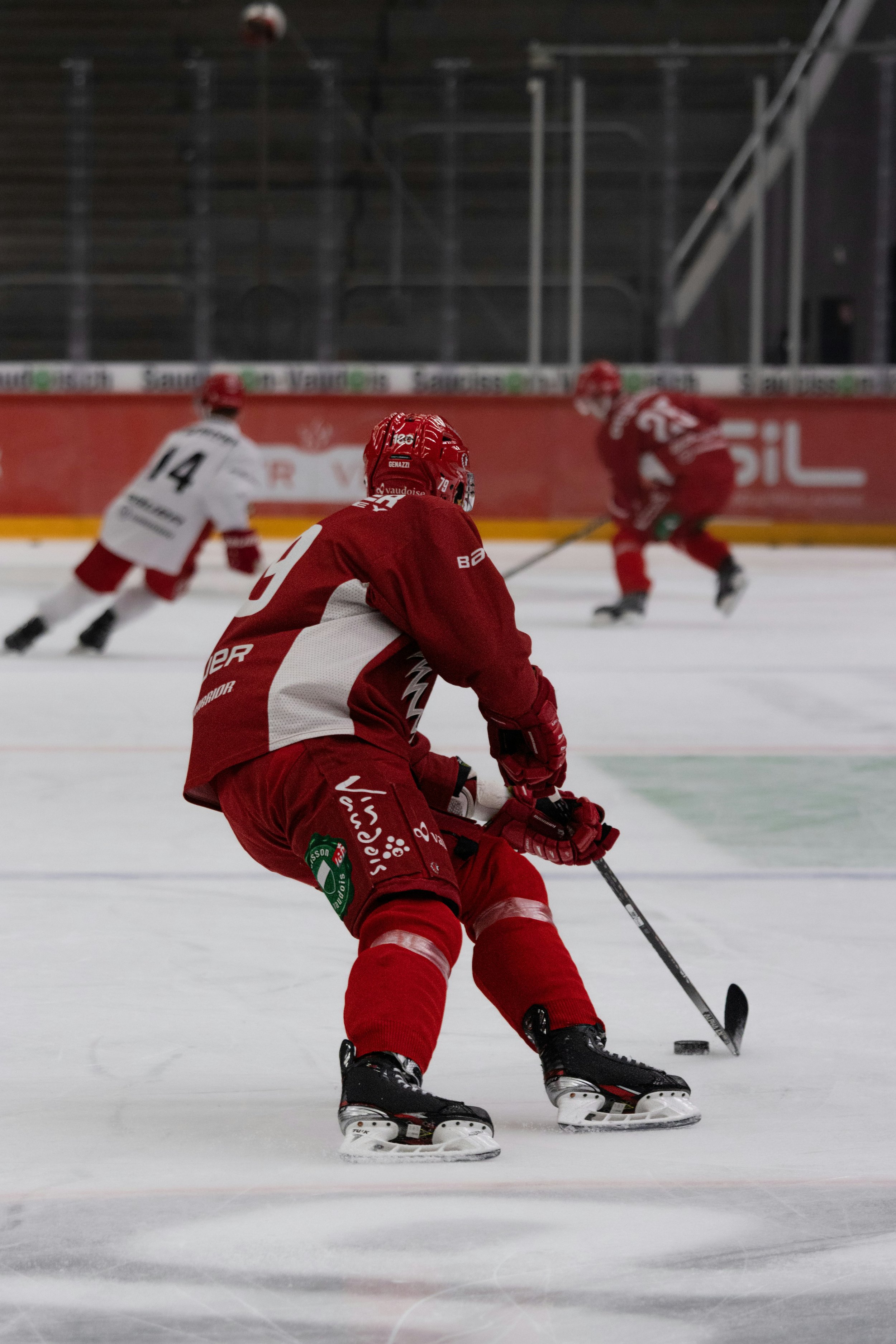 Ice hockey players in red and white uniforms on an indoor rink, with one player in the foreground handling the puck, and two other players in the background facing the ice.