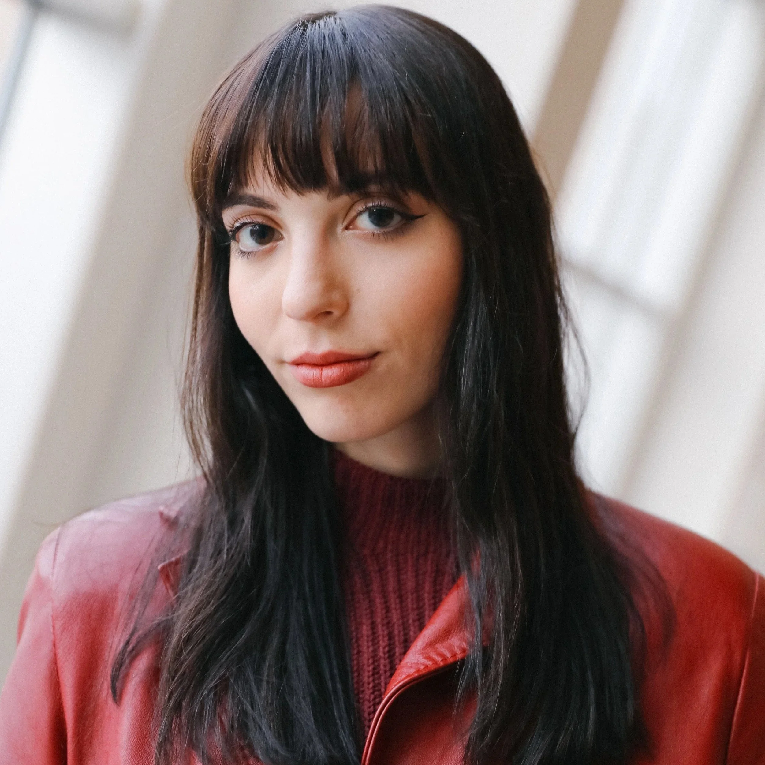 A woman with dark hair and bangs wearing a pink top and red leather jacket.