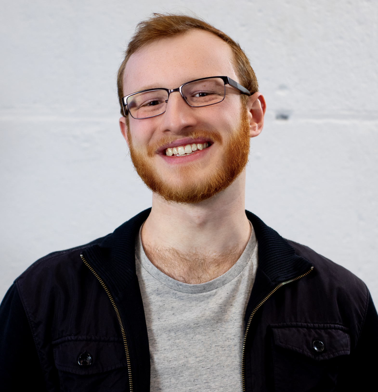 A young man with red hair and a beard, wearing glasses, a light gray t-shirt, and a black jacket, smiling against a plain light background.