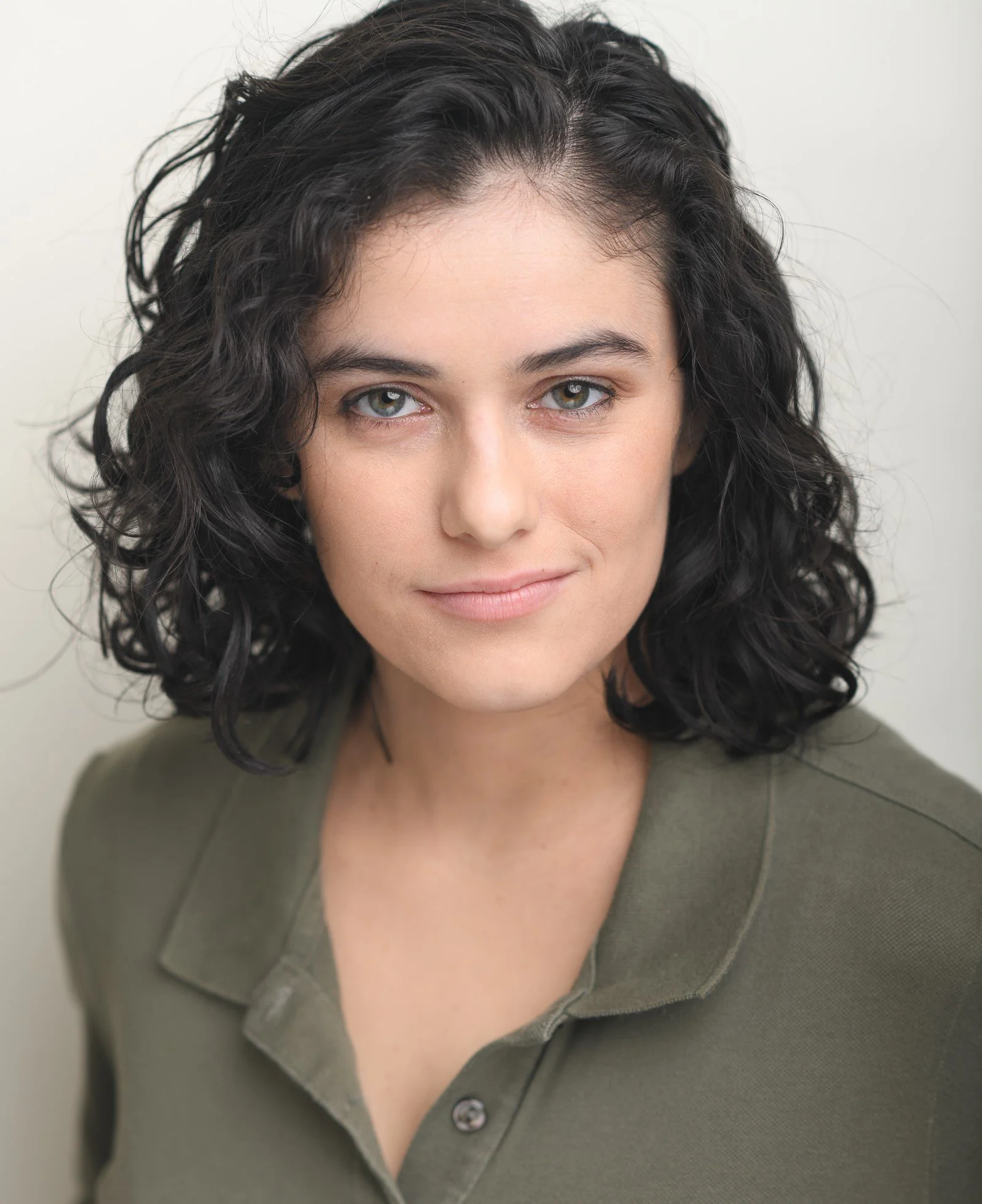 A woman with curly dark hair, wearing a green top with a collar, smiling subtly in front of a plain light background.