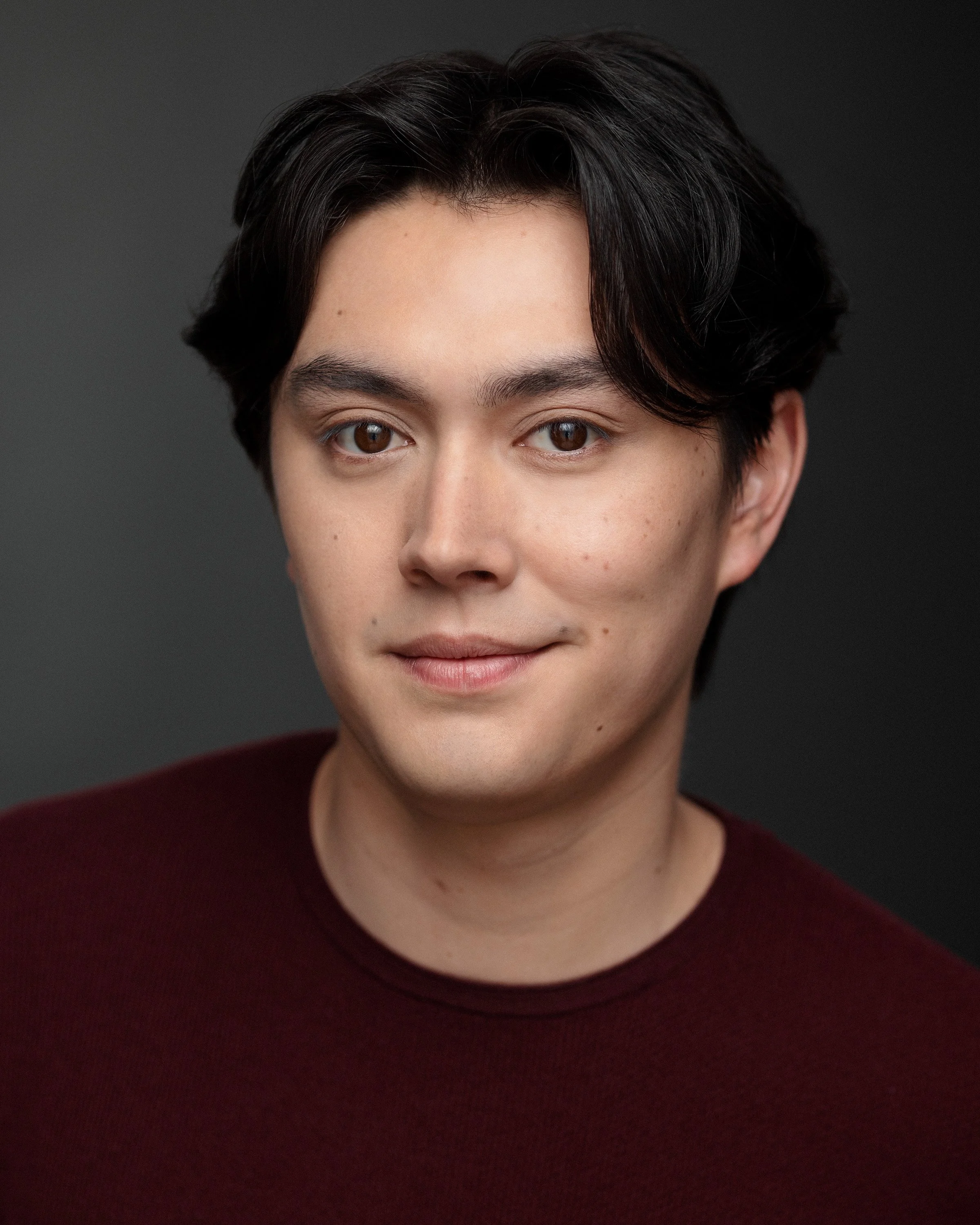 Close-up of a person with short dark hair, wearing a maroon top, against a gray background.