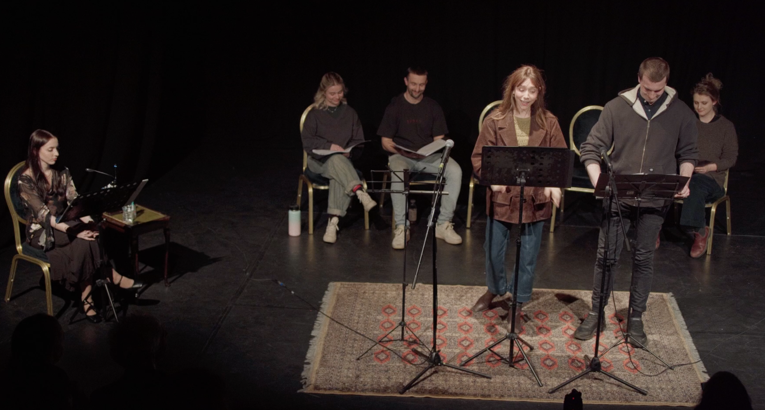 Group of people on a stage, some seated and some standing, engaged in a performance with microphones and music stands, set on a dark background with a patterned rug.