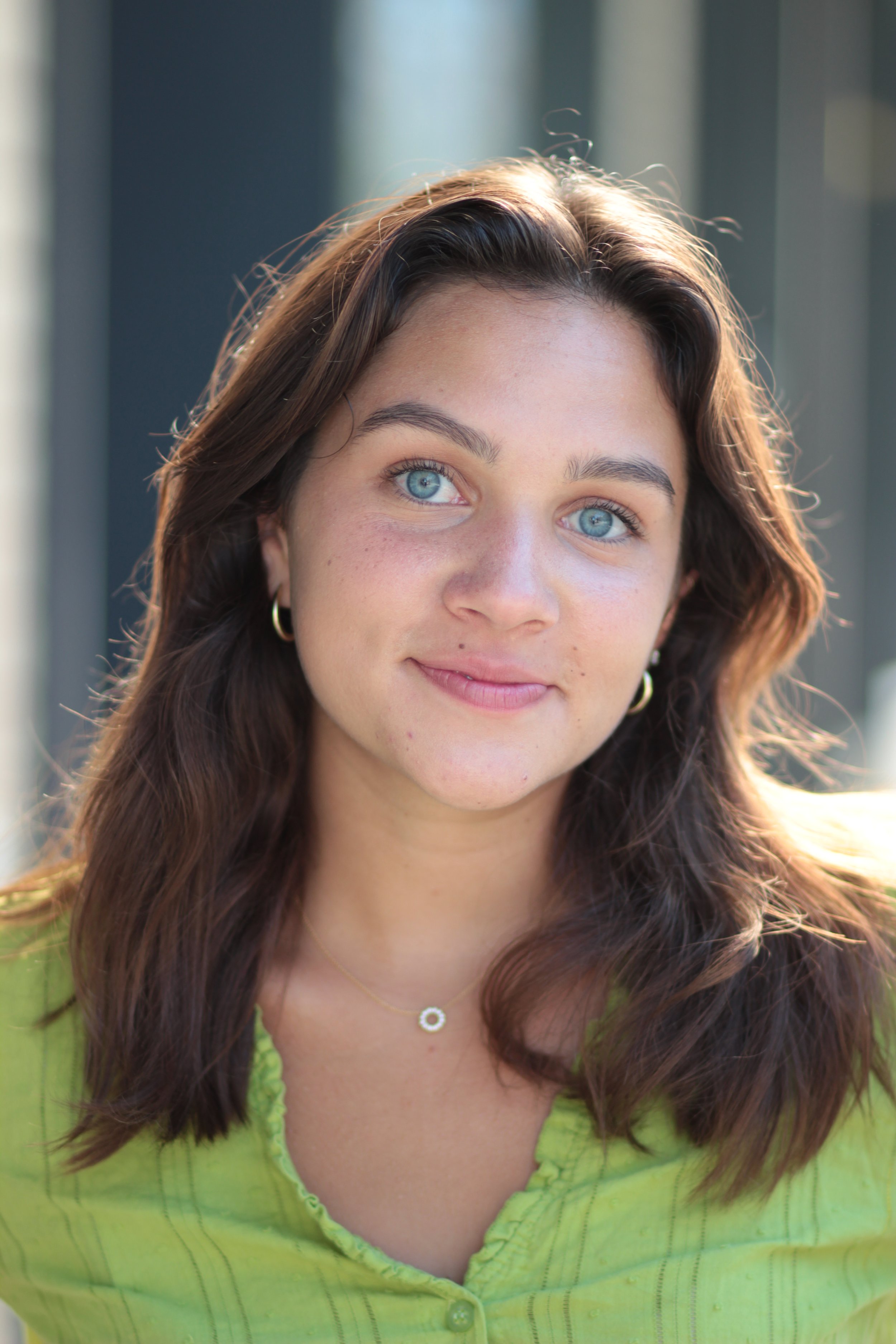 A woman with long brown hair, light skin, blue eyes, and wearing a green top and gold jewelry, smiling outdoors with blurred background.