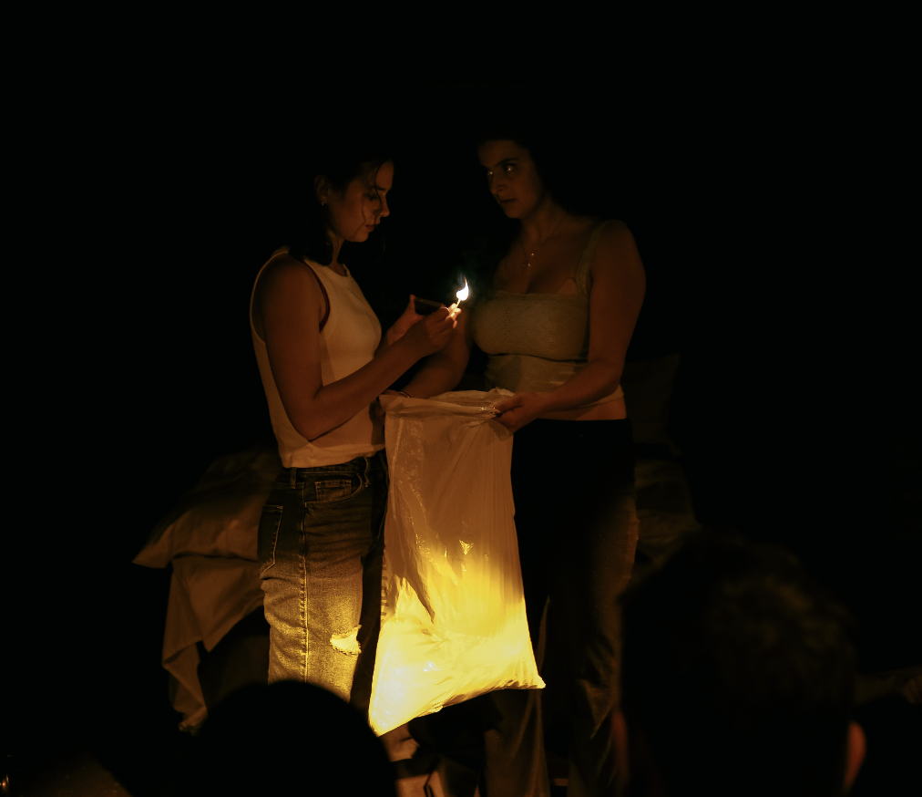 Two women in a dark room, one holding a match and the other holding a garbage filled with a glowing light, with a bed in the background.
