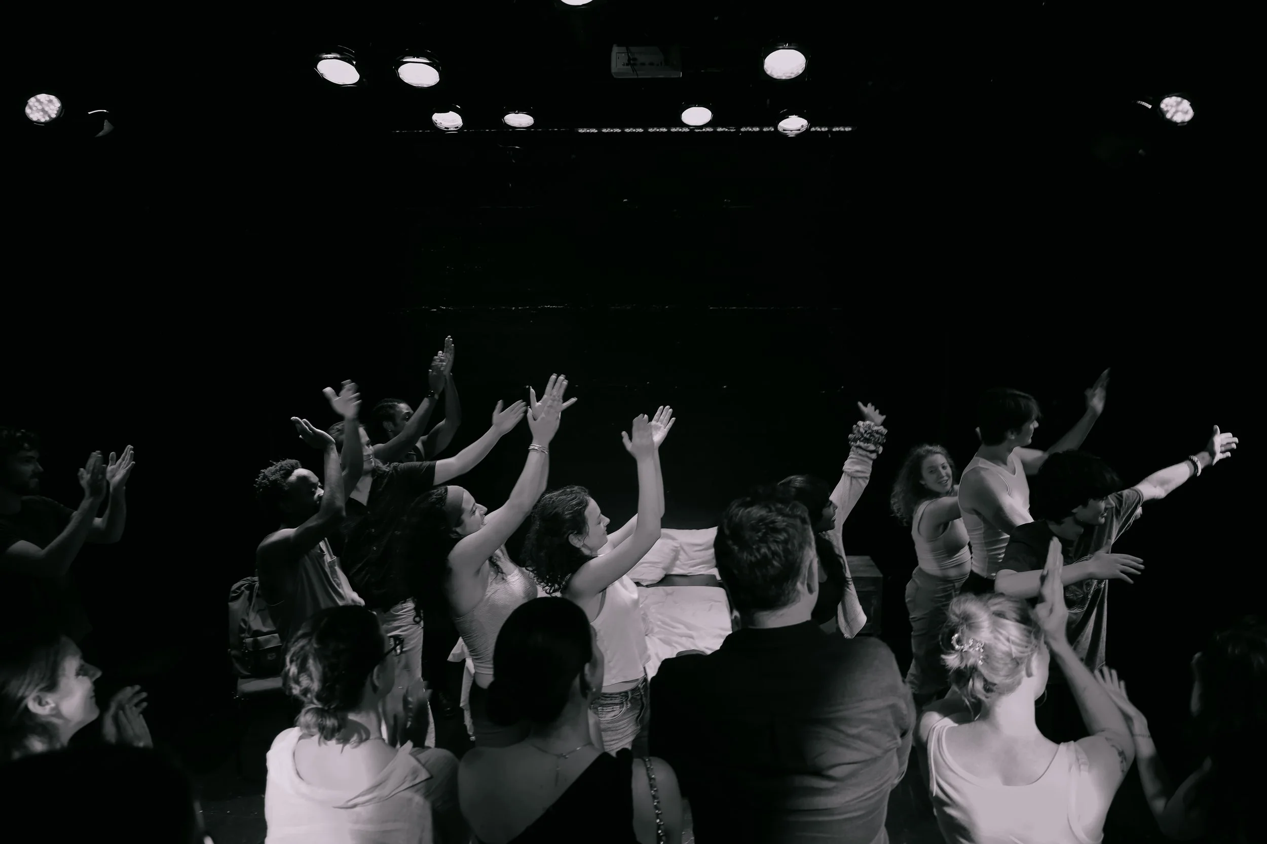 A group of people sitting and standing in a dark room with stage lights overhead, raising their hands and smiling.