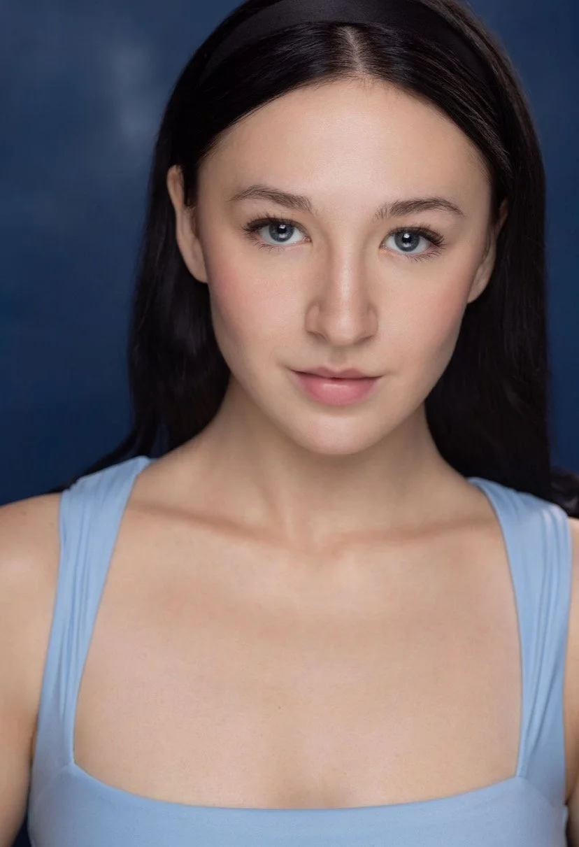 A young woman with long dark hair and blue eyes, wearing a light blue top, looking directly at the camera against a dark background.