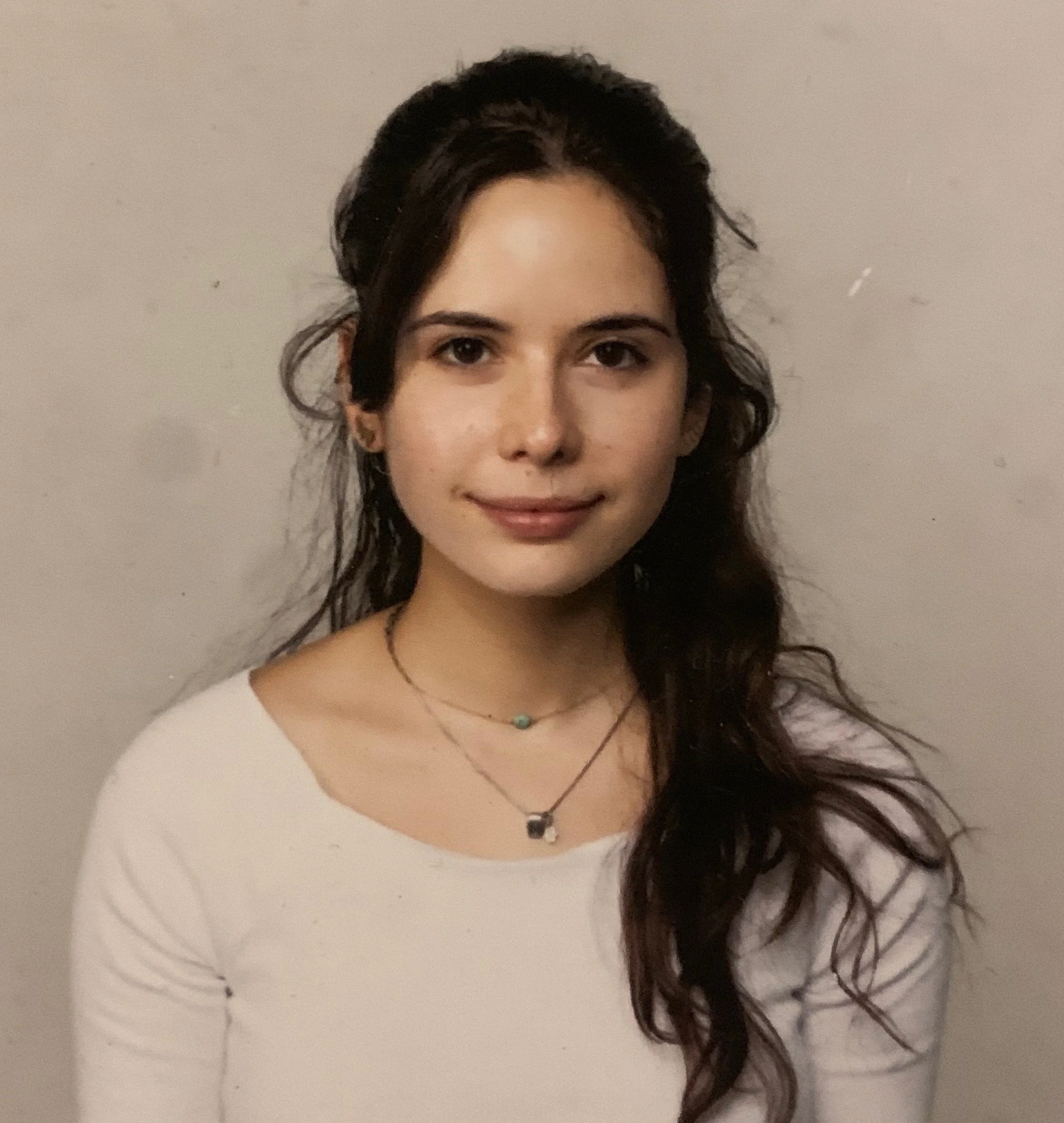 A young woman with dark, curly hair and light skin, smiling softly, wearing a white top and a necklace, standing against a plain wall.