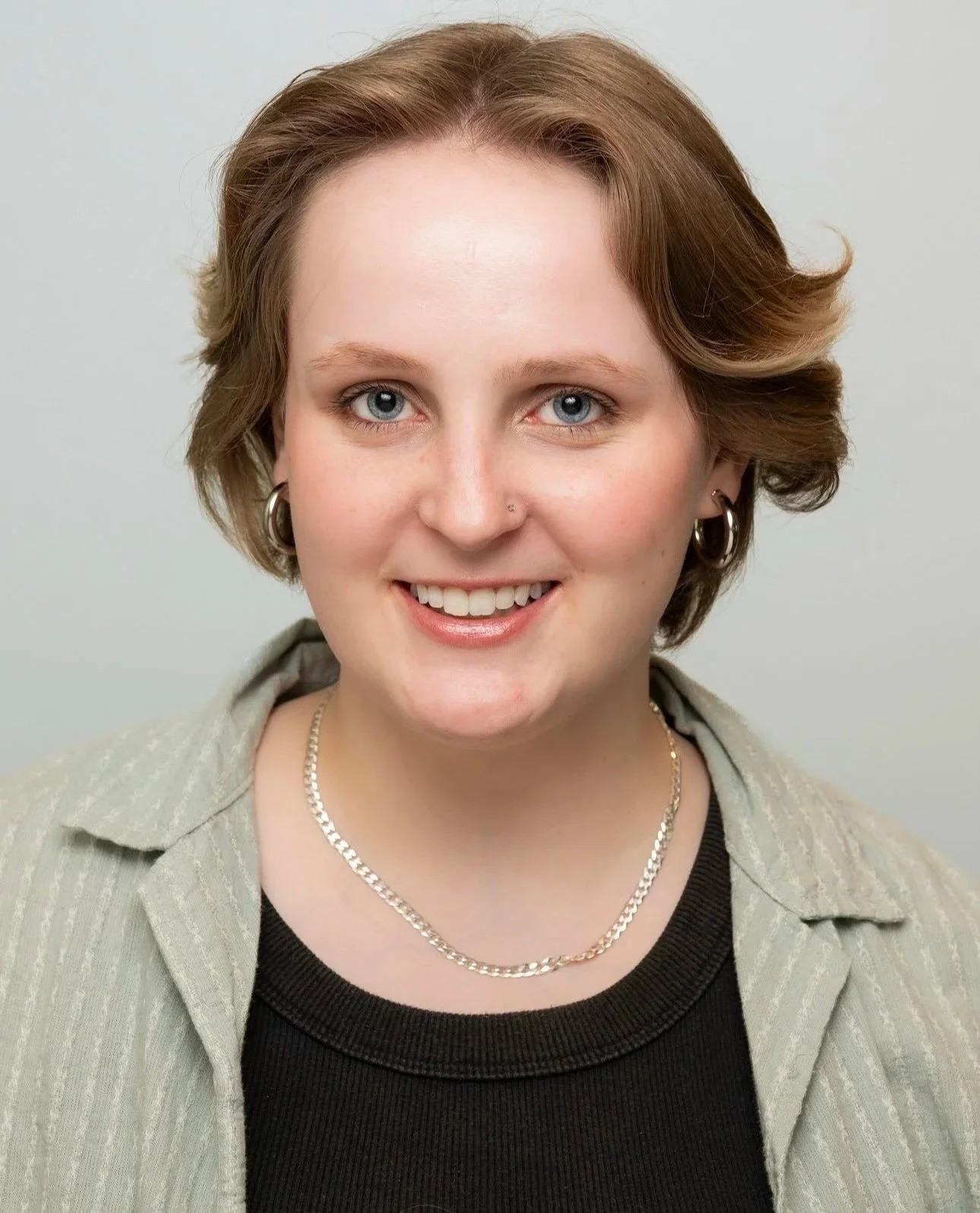A woman with short, light brown hair, blue eyes, and light skin smiling, wearing hoop earrings, a silver necklace, a black shirt, and a beige jacket against a neutral background.