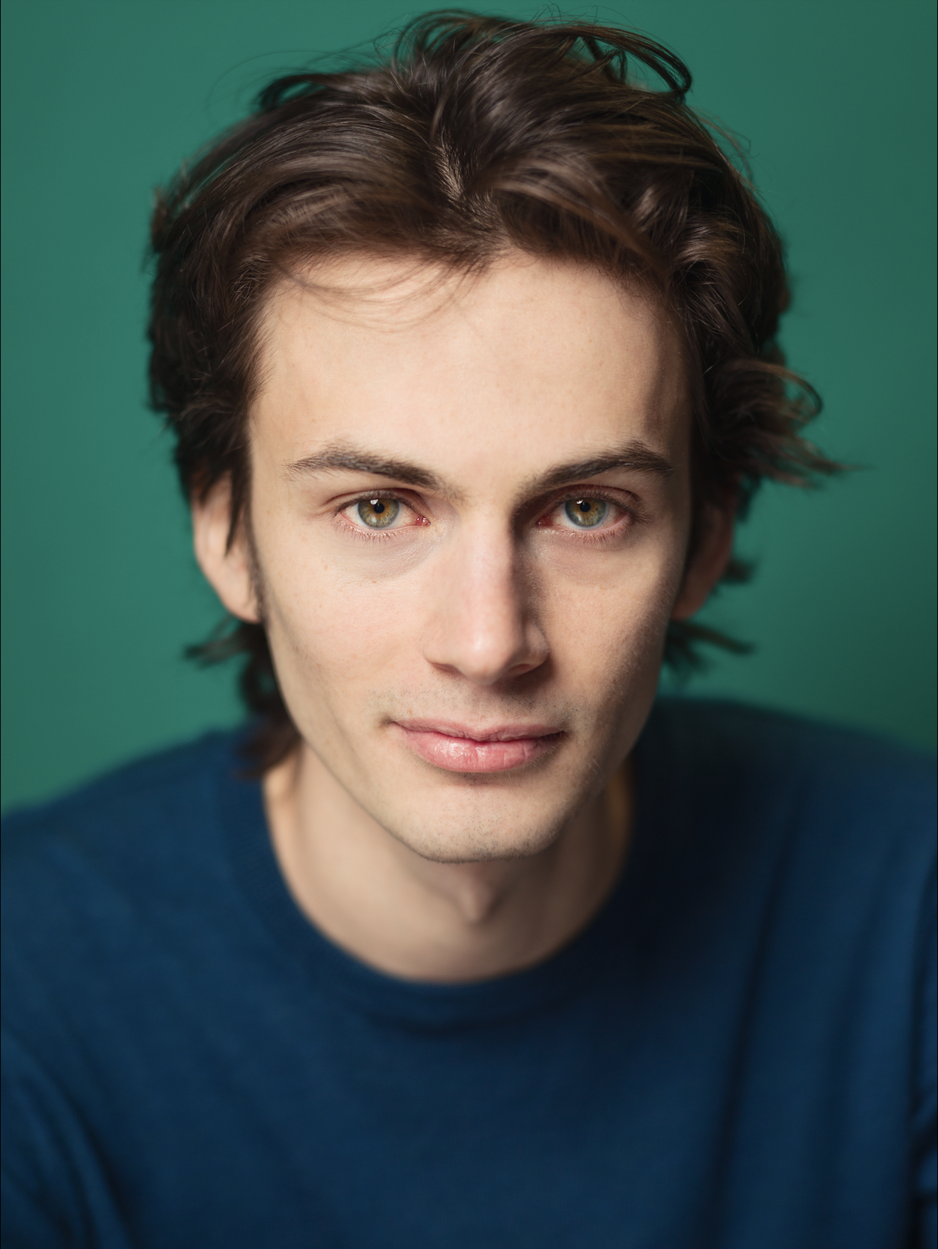 Close-up photo of a young man with brown, wavy hair, hazel eyes, and light skin, wearing a dark blue shirt, against a green background.