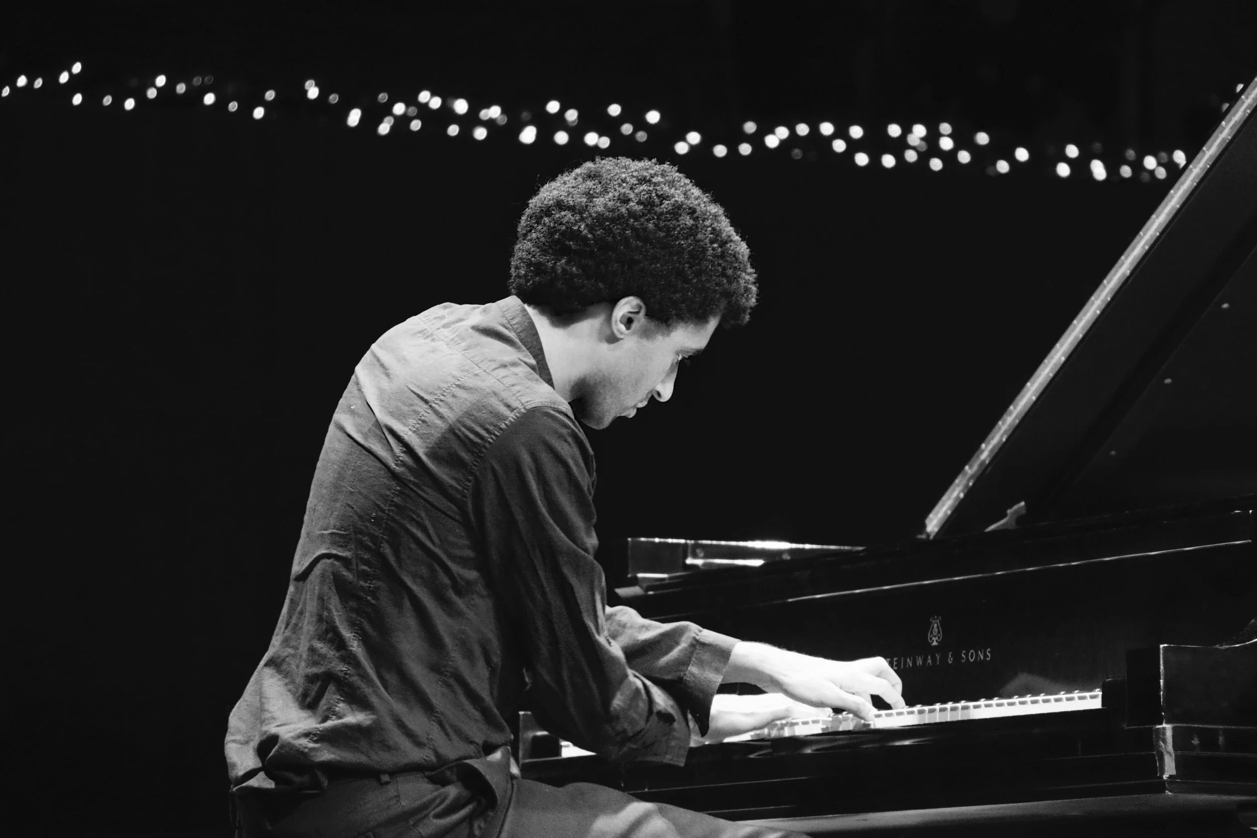 A person with short curly hair playing a grand piano in a dark setting, with a blurred string of lights in the background.