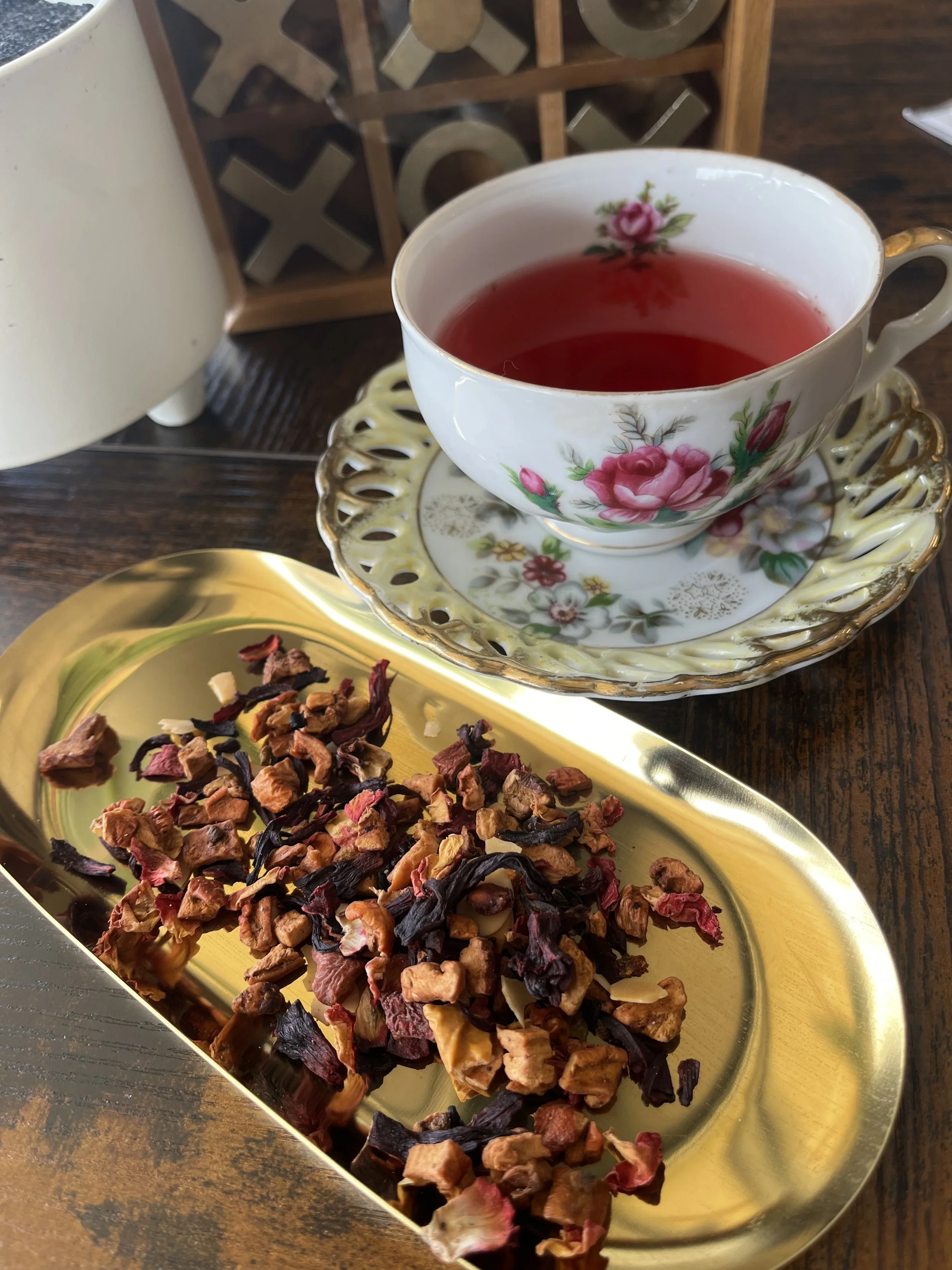A cup of red herbal tea with a floral pattern, placed on a matching saucer, and a gold tray with a mixed dried herbal tea blend on a wooden table.