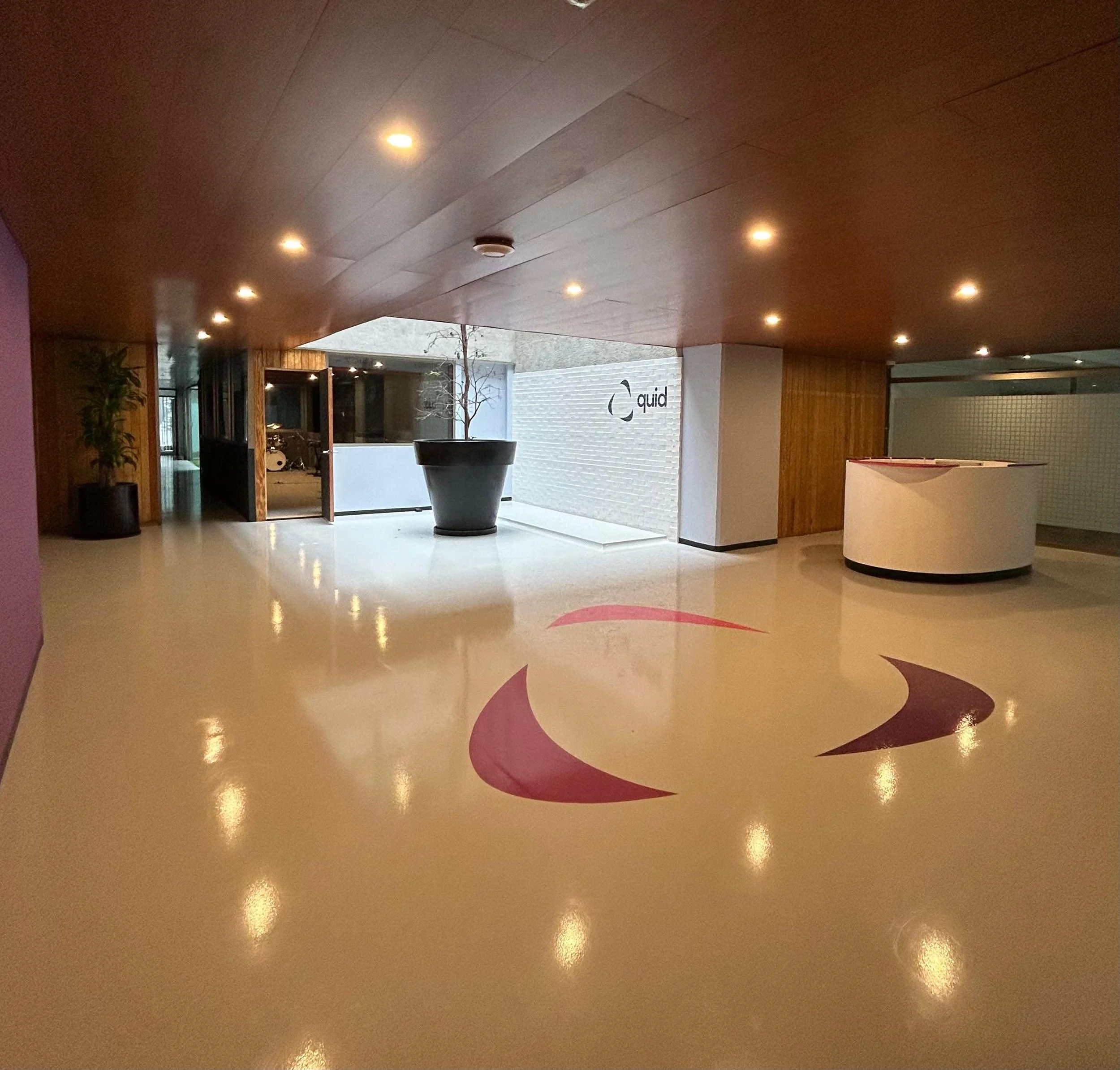 Interiores de un edificio con techo de madera y luces empotradas, paredes blancas y una pared con la palabra 'quid' y un símbolo en negro. El piso es de color beige con un diseño en tonos rosados y marrón.