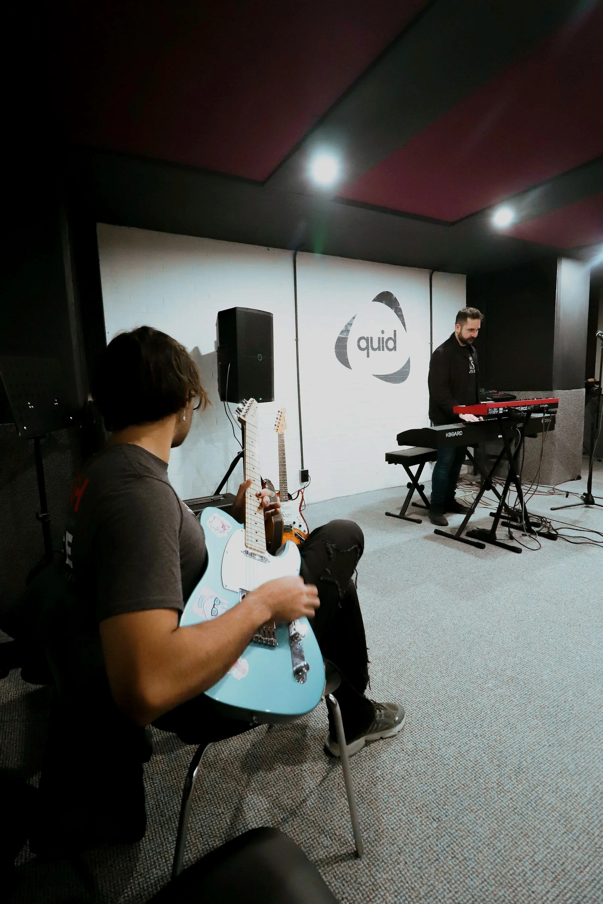 Una persona tocando guitarra y otra tocando teclado en una sala de música, con un logo de 'quid' en la pared de fondo.