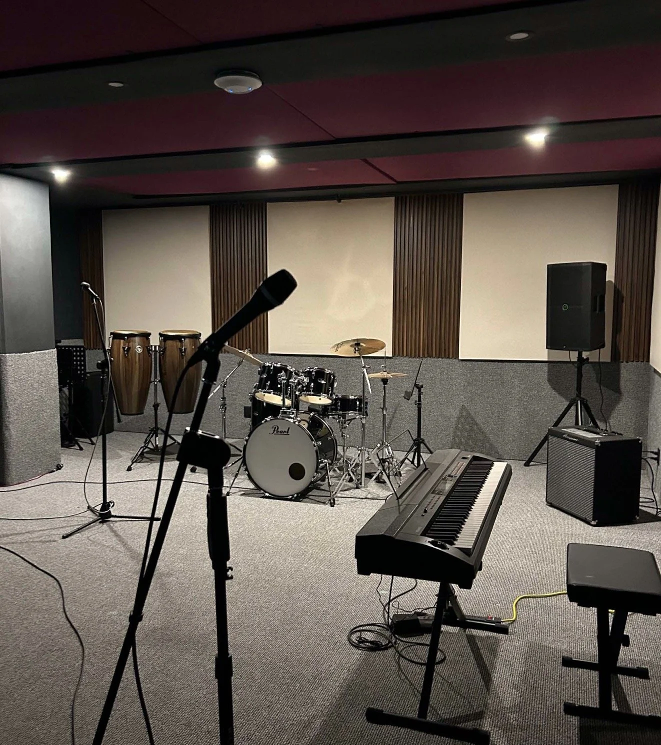 Un escenario de música con instrumentos incluyendo batería, teclado, micrófono, conactos, y altavoces en una sala de ensayo o presentación.