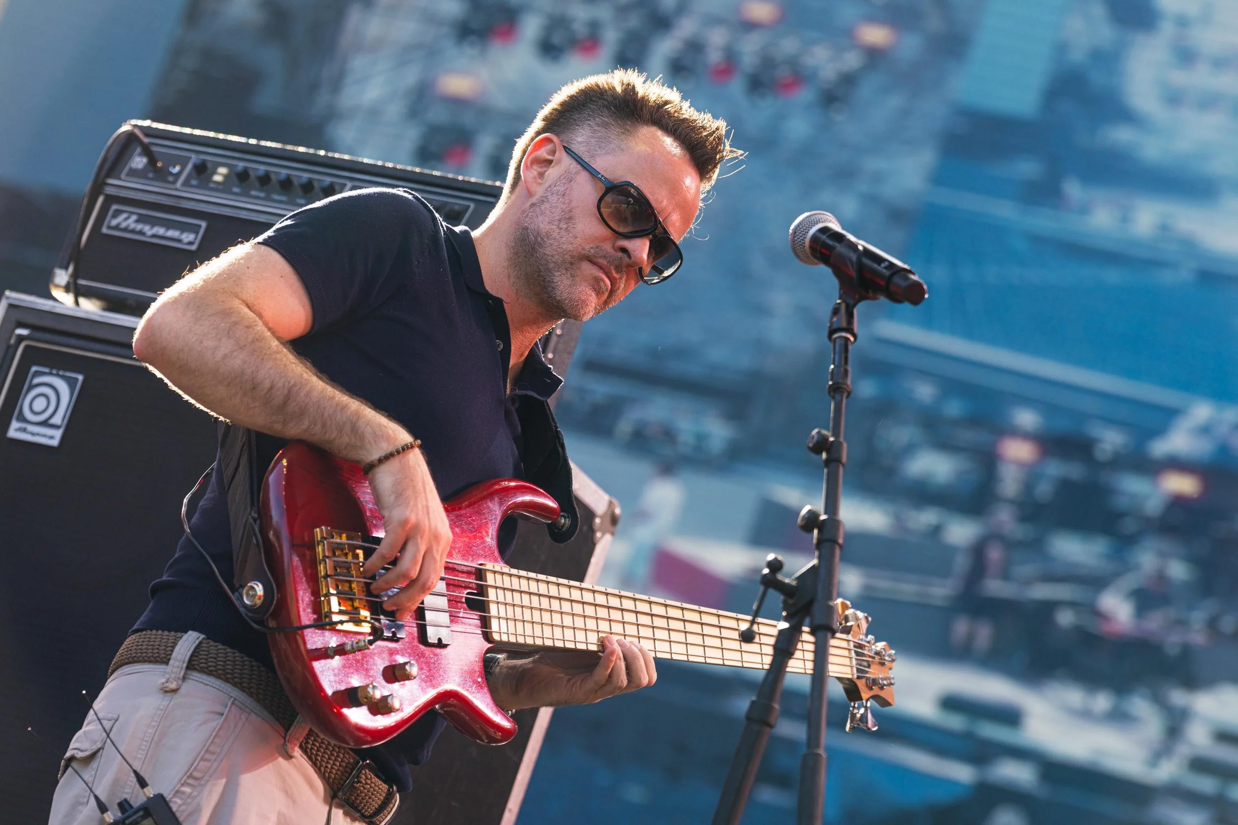 Un hombre tocando una guitarra eléctrica en un escenario al aire libre, con amplificadores y una ciudad de fondo.