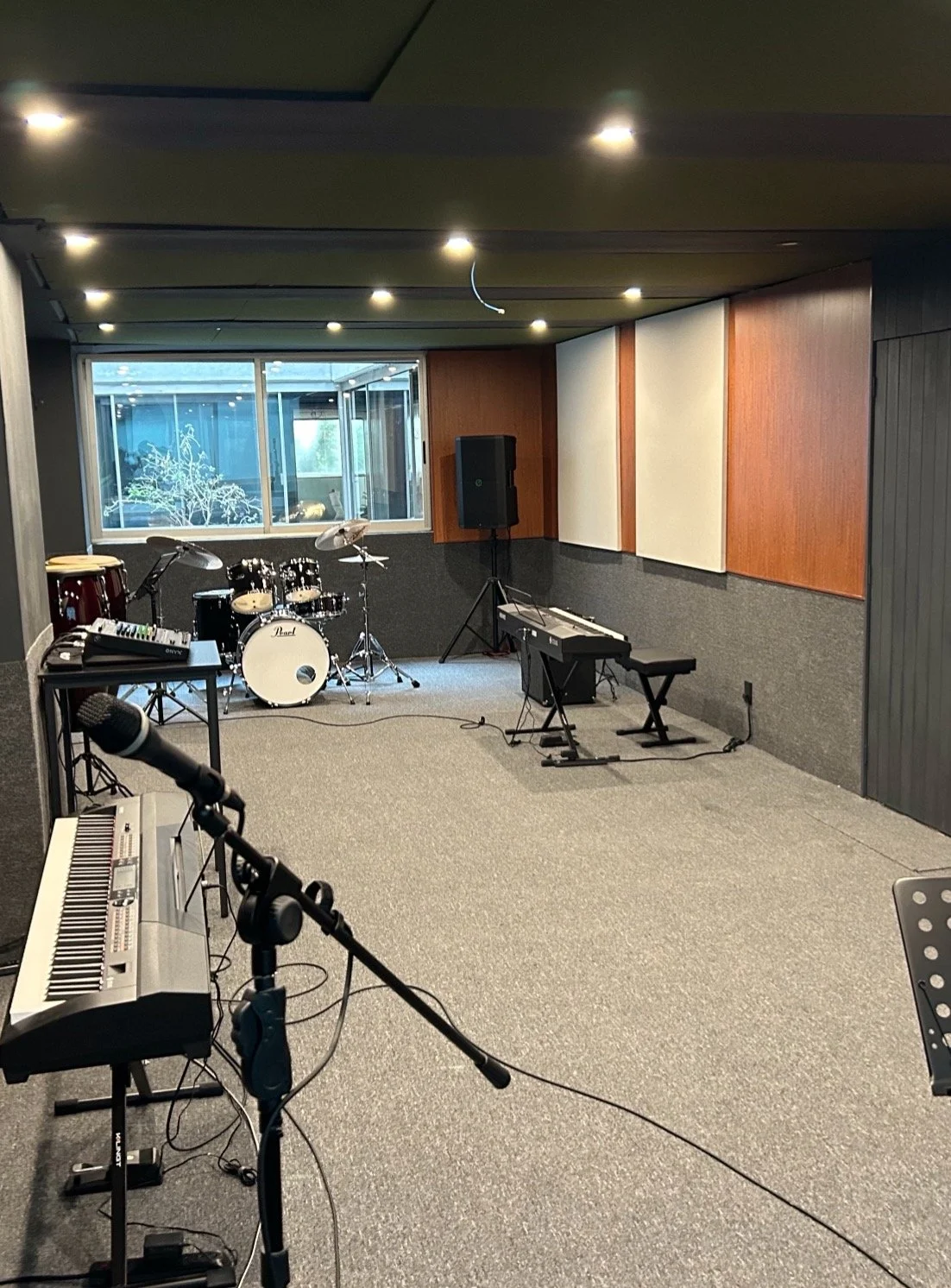 Sala de ensayo con batería, teclado, micrófono, sistema de sonido y paneles acústicos en las paredes.