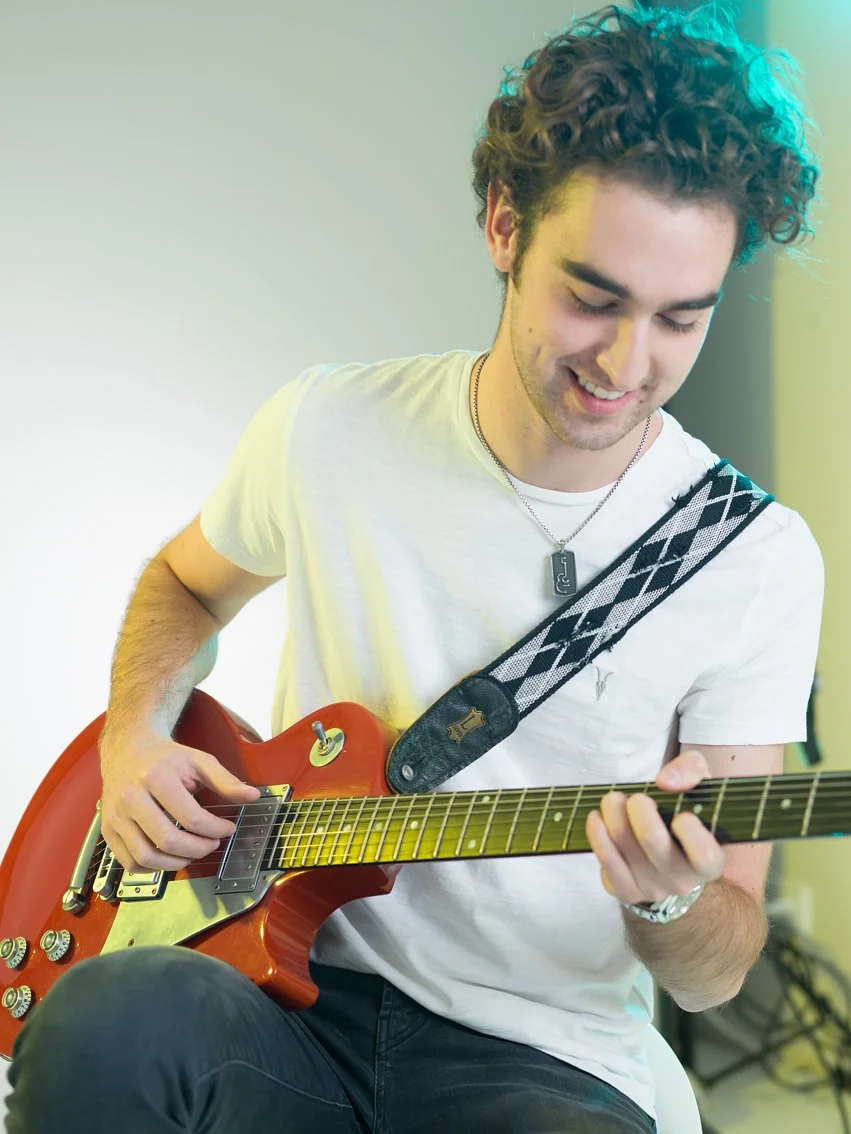 Joven tocando guitarra eléctrica de color rojo, sentado, sonriendo, en un ambiente con iluminación suave.