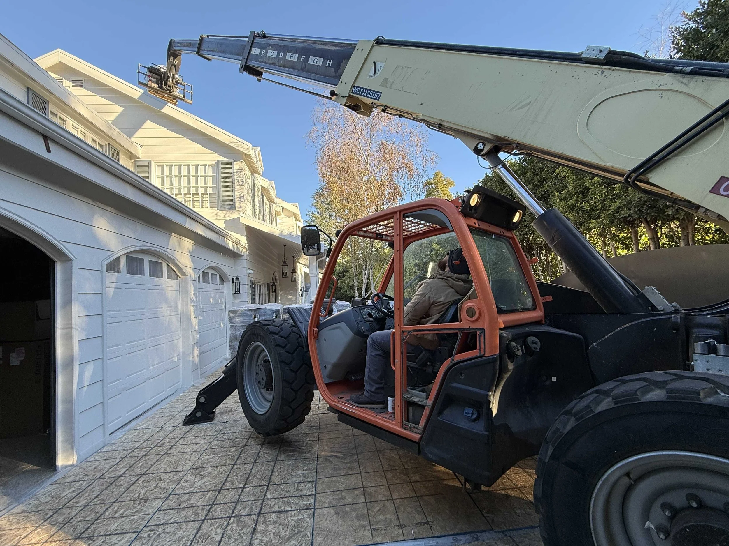 A person operating a construction lift truck outside a white residential building with a garage, trees, and blue sky in the background.