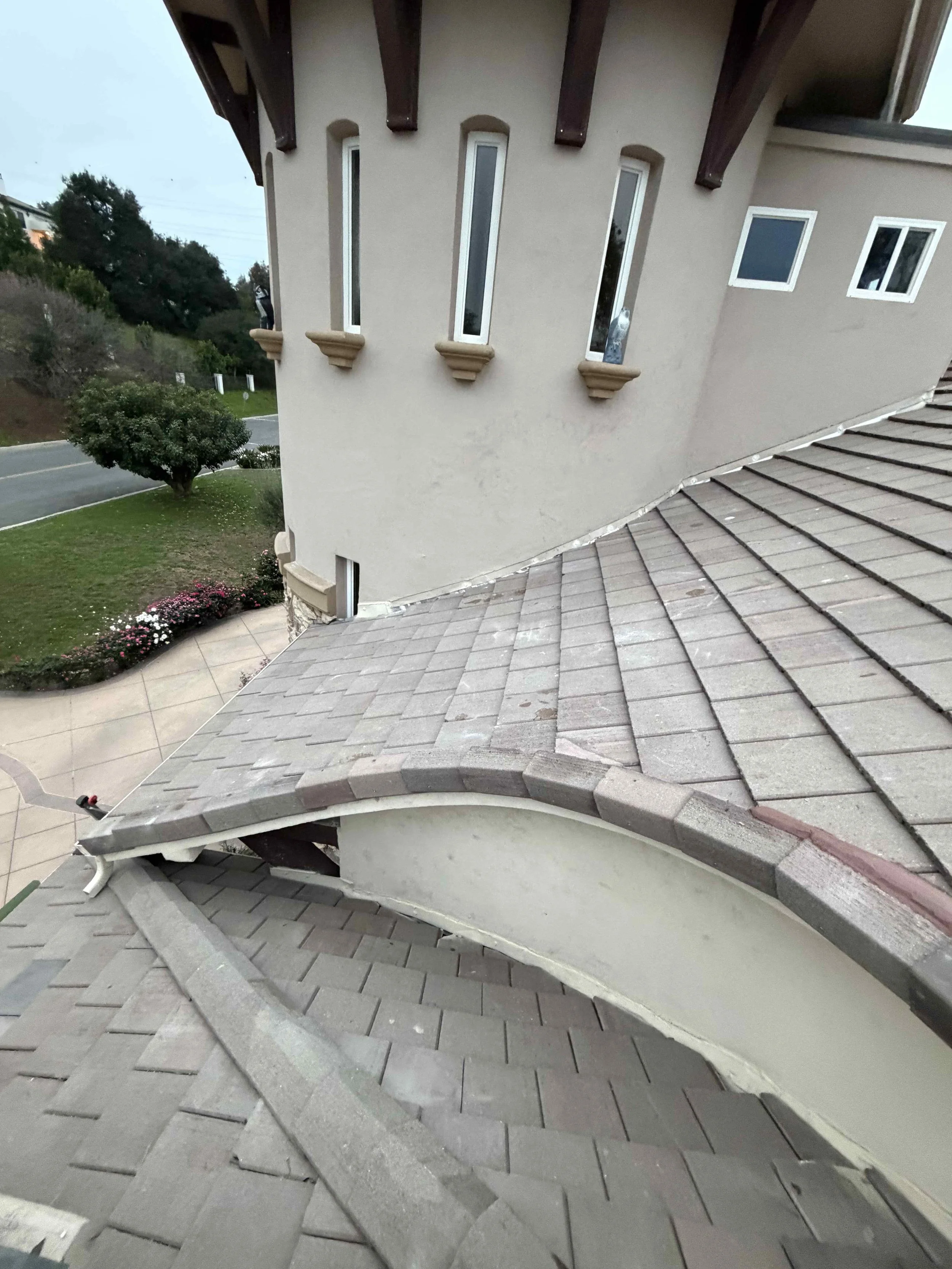 View of a rooftop with damaged shingles and missing tiles, part of a beige-colored house with decorative window ledges, adjacent to a landscaped yard with flowers and trees.