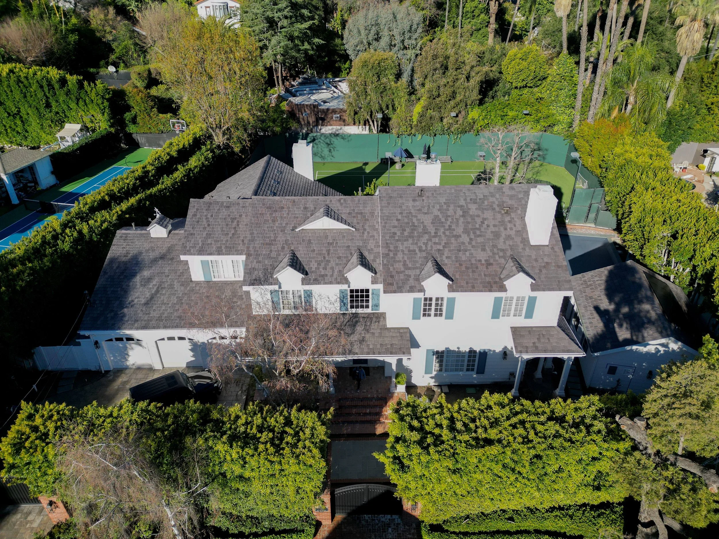 An aerial view of a large house with gray roof, surrounded by tall trees, a backyard with a tennis and basketball court, and lush green landscaping.