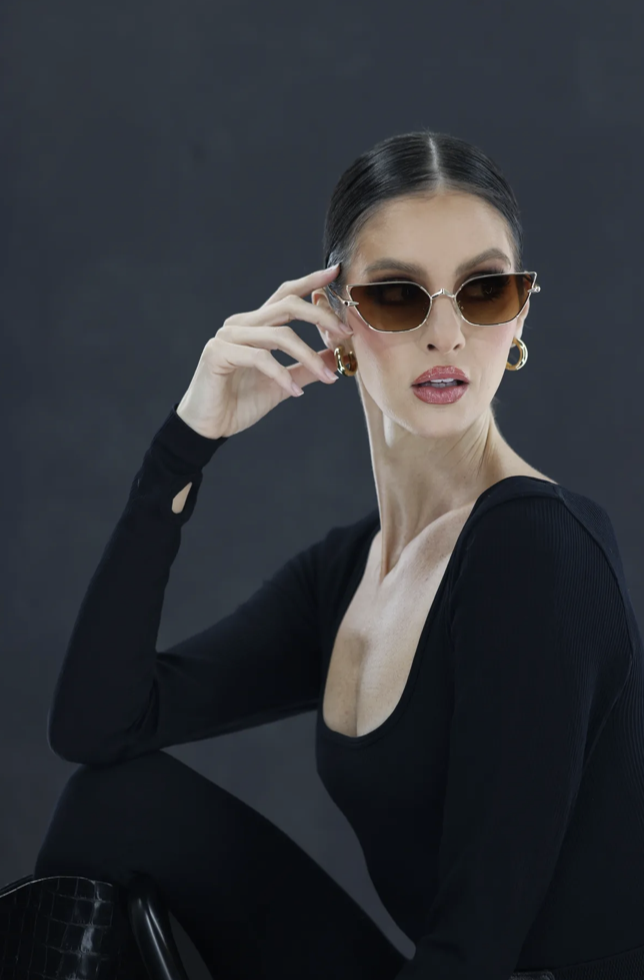A woman with dark hair slicked back, wearing sunglasses, gold hoop earrings, a black long-sleeve top, and black pants, posing against a dark background.
