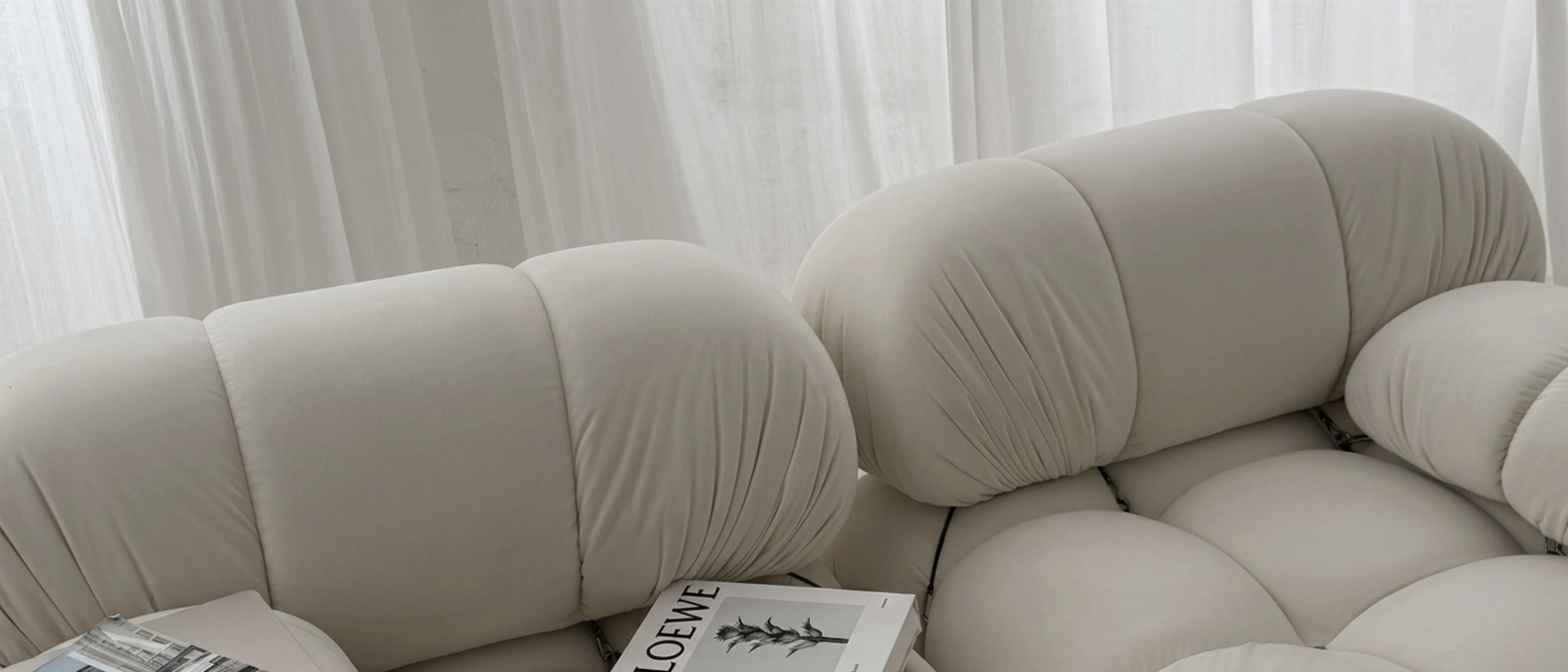 Close-up of a white, plush, tufted sofa with a magazine resting on it, in front of sheer white curtains.