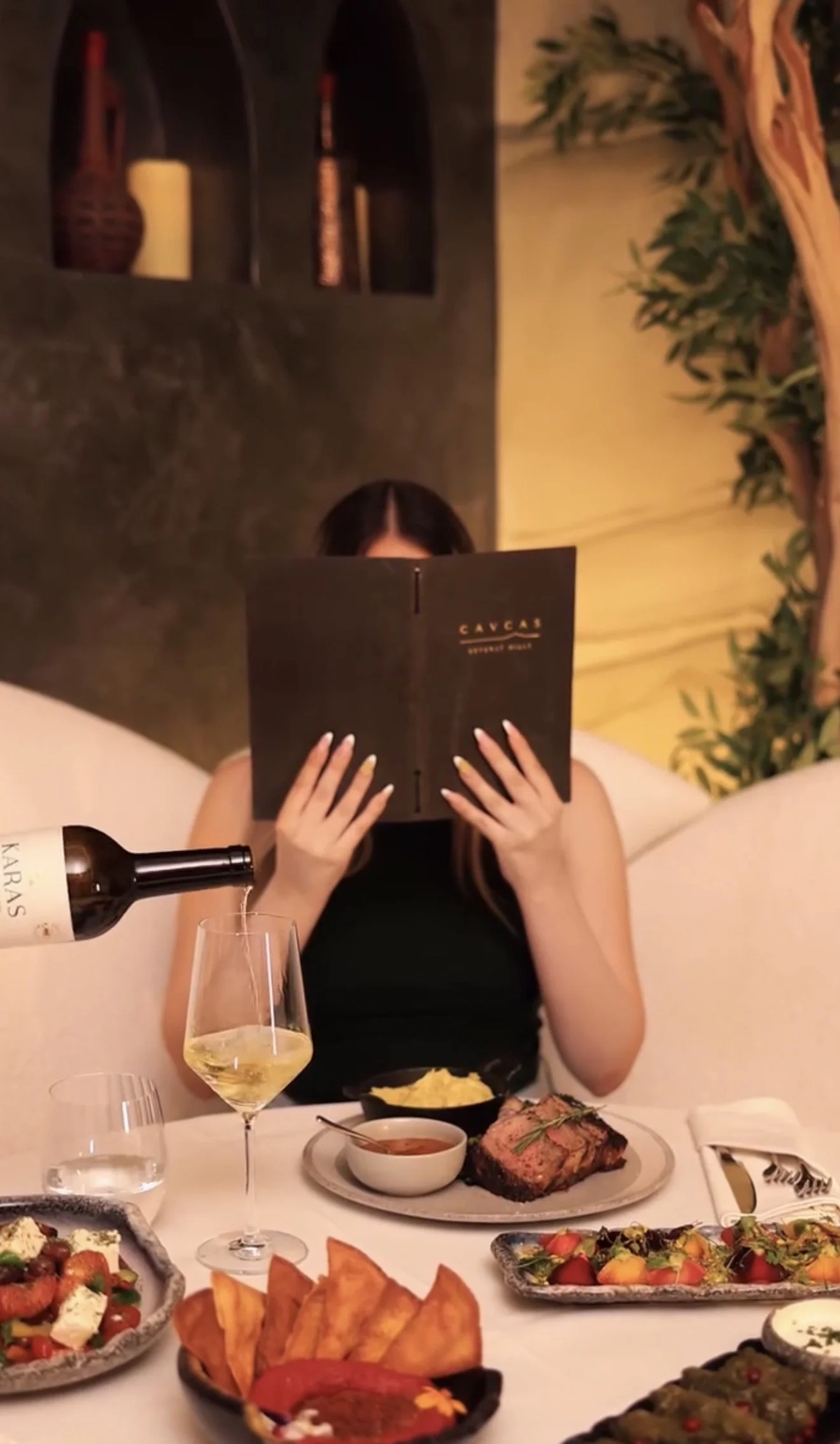 A woman dining at a restaurant, reading a menu titled 'Cavas'. She is holding a menu in front of her face. On the table, there is a bottle of white wine being poured into a glass, a glass of water, and various dishes including a steak, salads, potato chips, and other appetizers.
