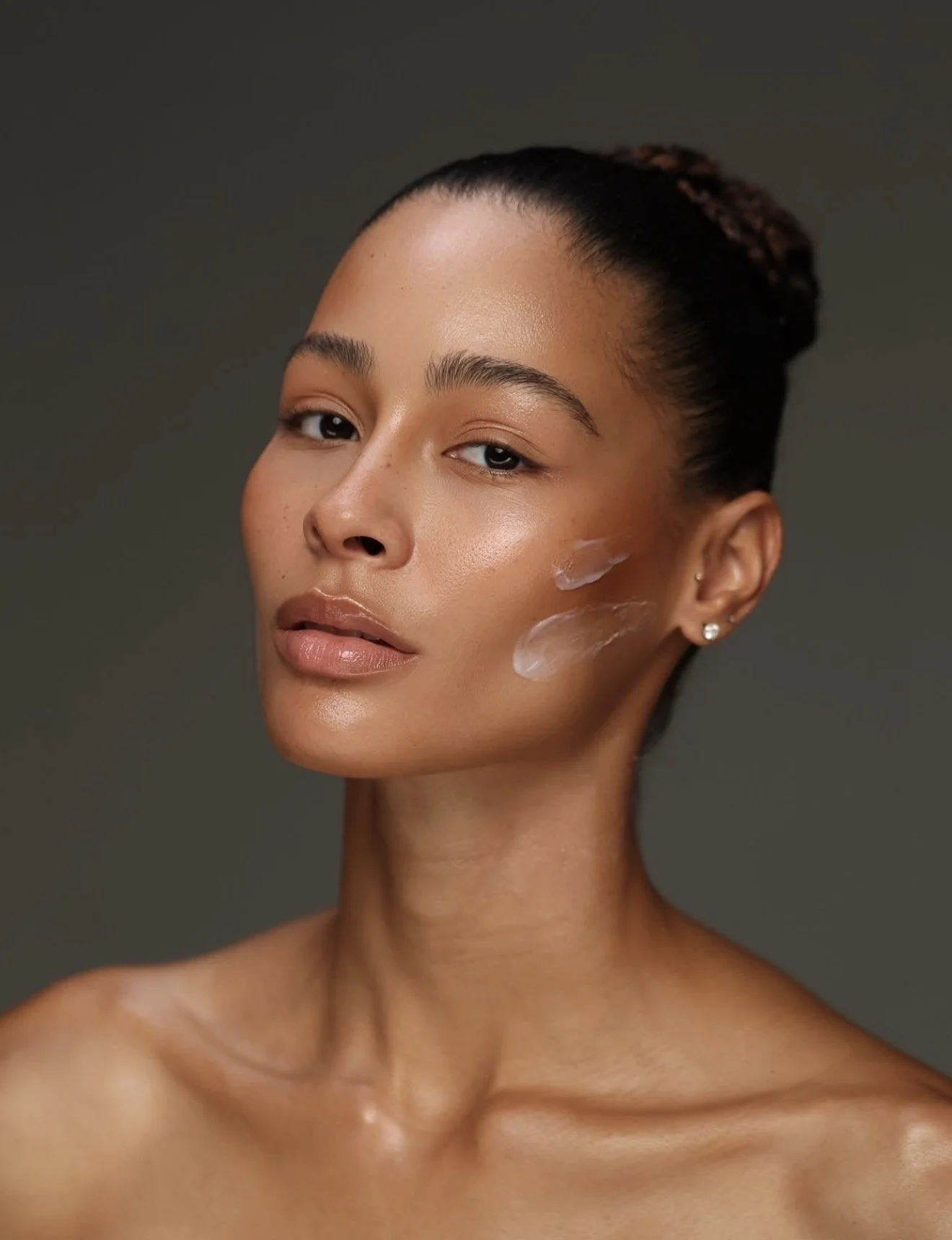 Close-up of a woman with glowing skin and two streaks of skincare product on her cheek, looking directly at the camera with her hair in a bun.
