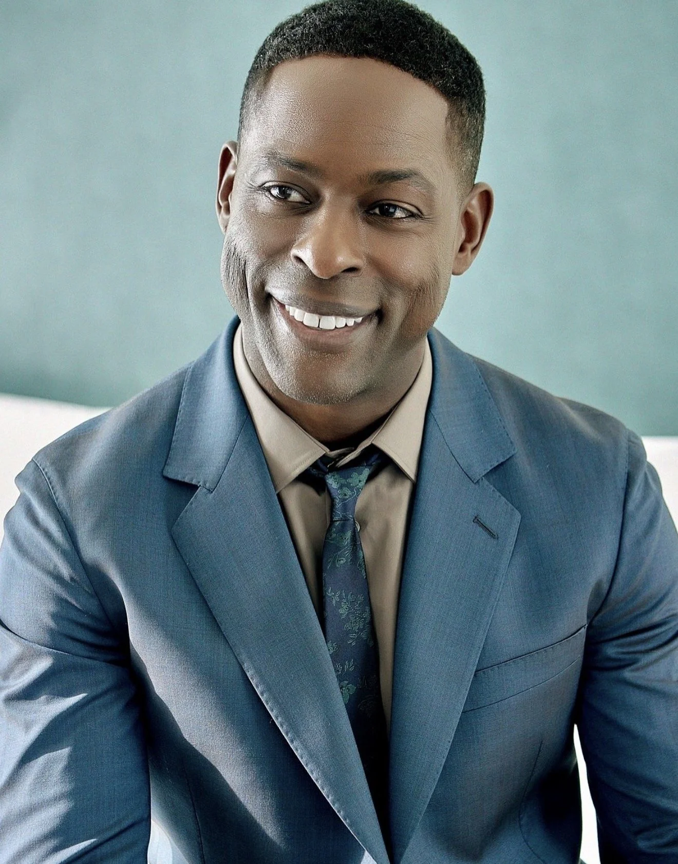 A smiling Black man wearing a blue suit, beige shirt, and patterned tie, sitting in front of a light teal background.