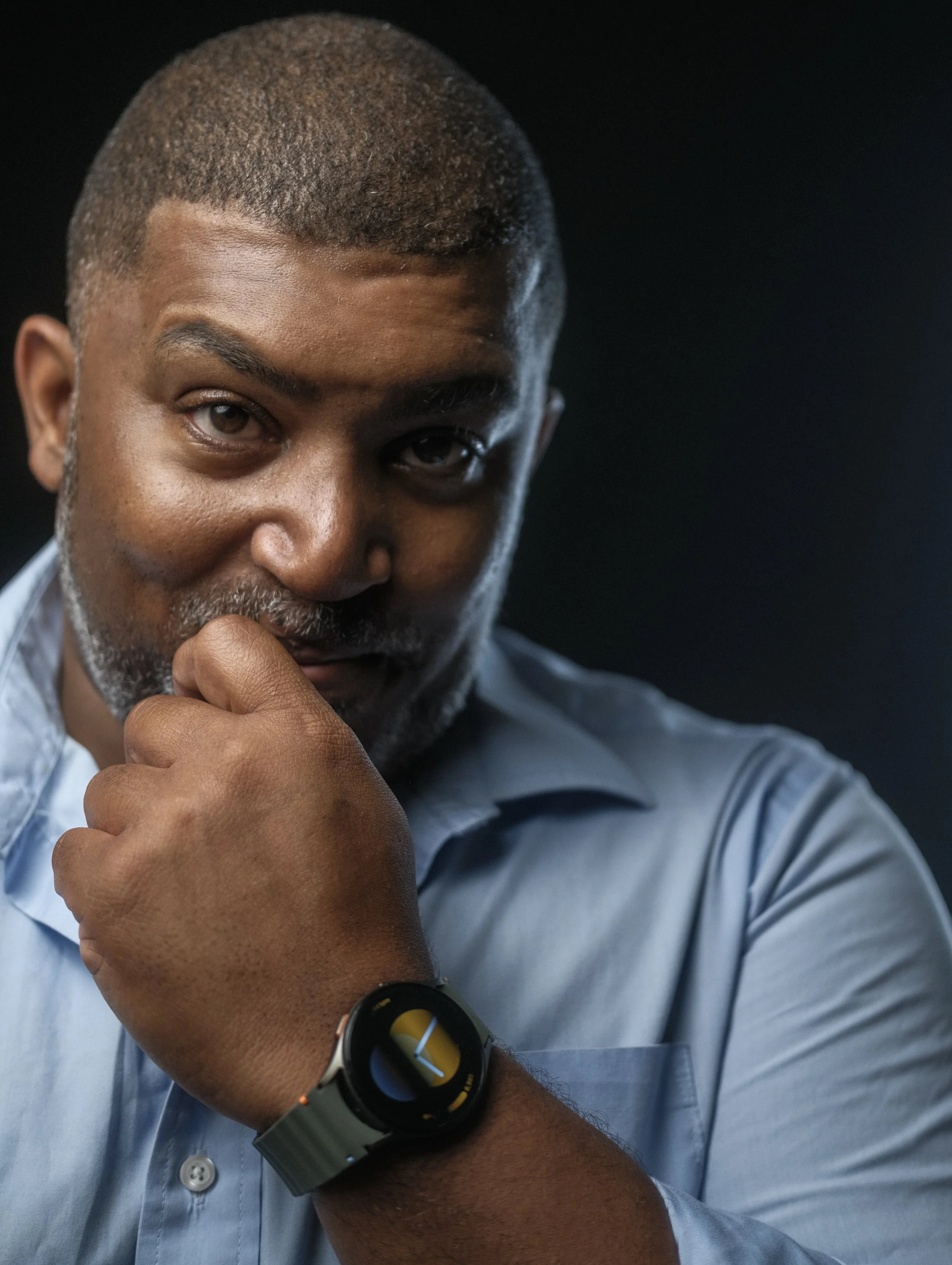 A man with short hair and a beard wearing a light blue shirt, looking thoughtfully at the camera with his hand near his chin, wearing a smartwatch on his wrist.