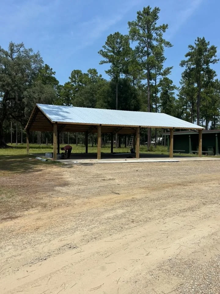 A wooden picnic shelter with a metal roof in an outdoor park area, with a person working on construction and surrounded by tall trees.