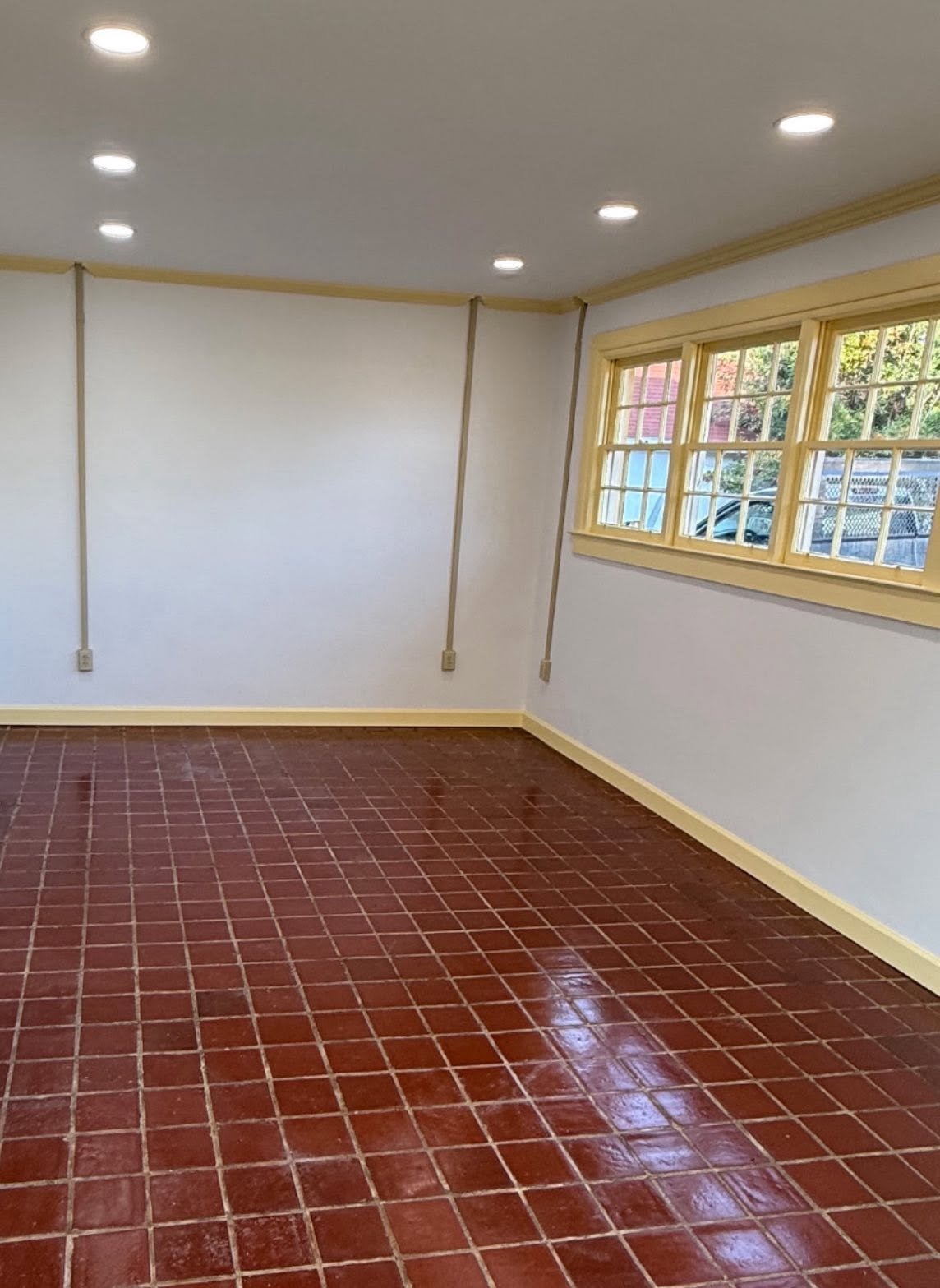 Empty room with white walls, yellow trim, red tile floor, and multiple ceiling lights; a window with yellow trim shows outdoor greenery.